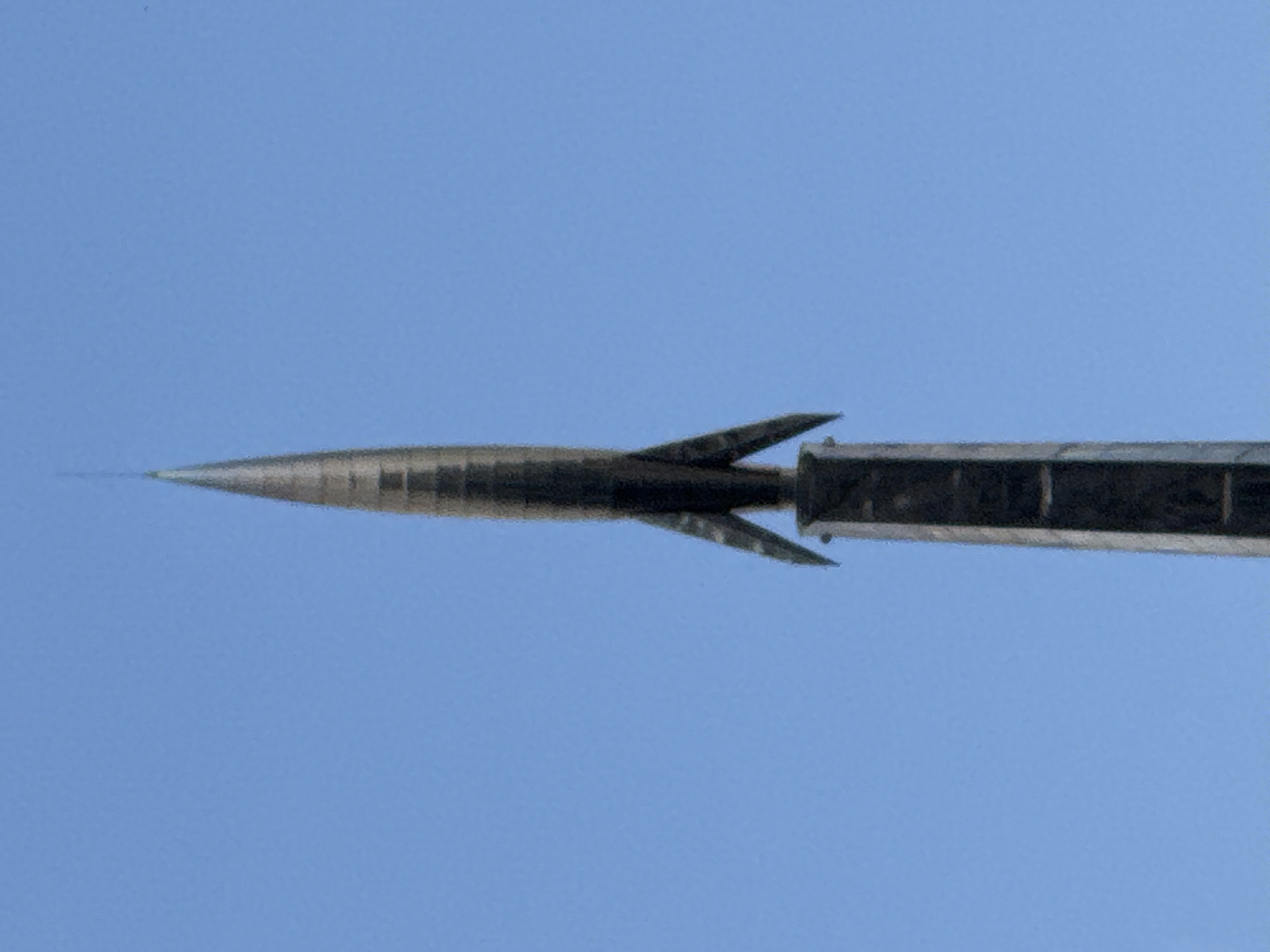 The image displays a metallic object, possibly a missile or a dart, against a clear blue sky. The object is elongated and pointed at the tip, with fins extending from its rear. It appears to be in motion or poised for launch. The object's surface has a textured, segmented appearance, suggesting it's made of multiple components. The sky is uniformly blue, indicating daytime with fair weather. There are no discernible people, animals, or any other elements in the scene besides the object and the sky. The image has a slightly grainy texture, which may be due to the camera or resolution. The composition focuses solely on the object, with no background context apart from the sky. The time of day is likely during daylight hours. The location context provided is Moscow, Russia, but there are no specific visual cues within the image to confirm this. No text is visible in the image.