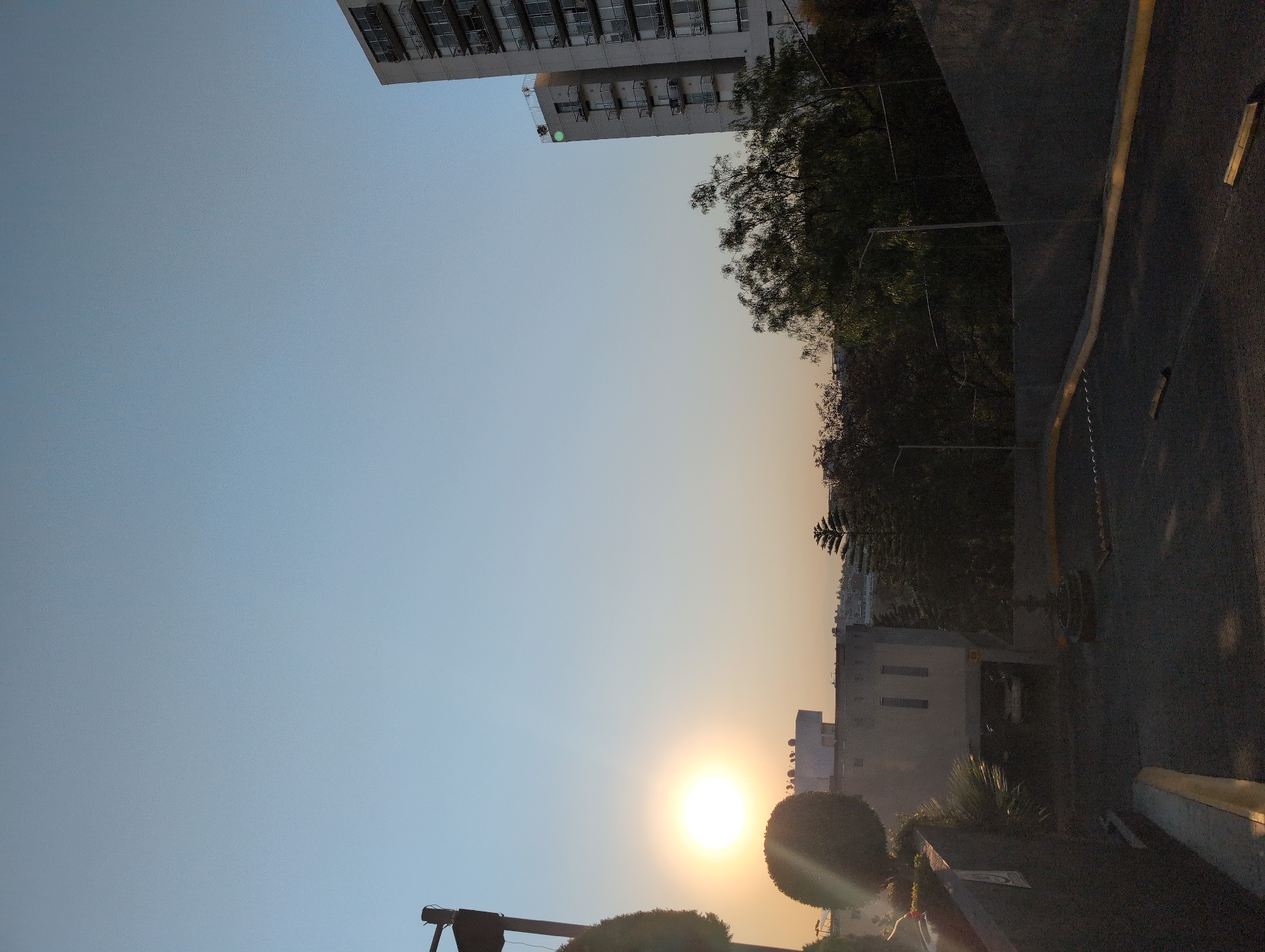 The image depicts a street scene at sunset in Huixquilucan de Degollado, Mexico. The sun is low on the horizon, creating a warm, golden glow in the sky. A tall apartment building is visible in the upper left corner. Trees line the street, and a building is visible in the middle ground. A street with a curb is on the right side of the image.
