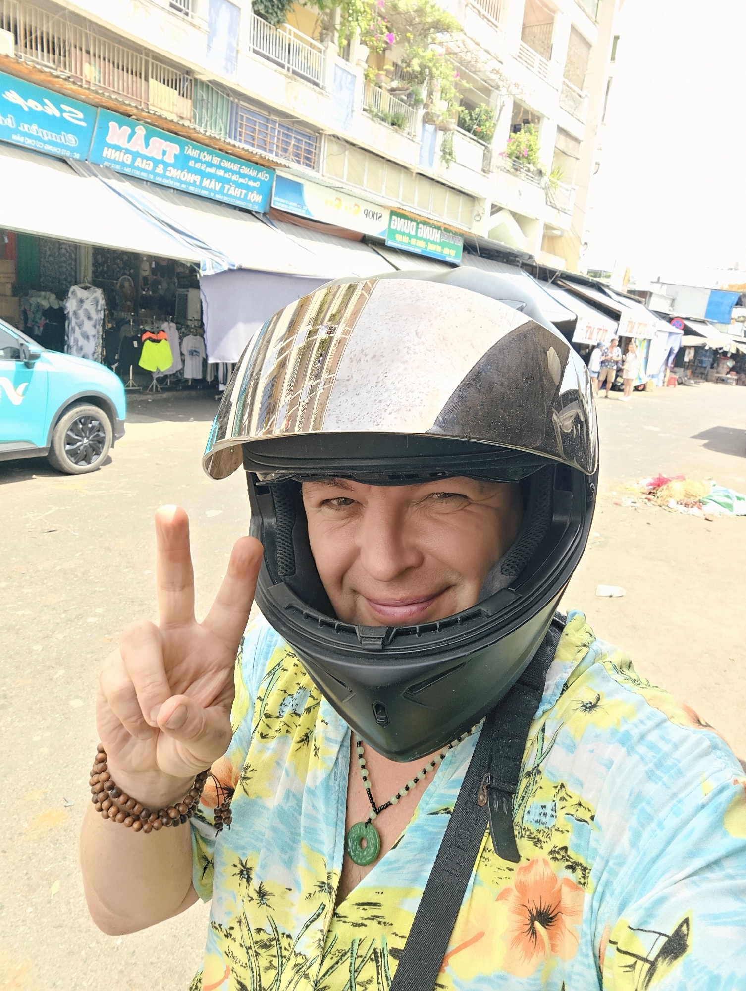 A person wearing a motorcycle helmet and a colorful tropical shirt is taking a selfie on a street in Nha Trang Ward, Vietnam. The person is smiling and holding up two fingers in a peace sign. In the background, there are shops with awnings, a turquoise car, and other people. The street appears to be a market area with stalls and merchandise. The buildings in the background are multi-story structures with balconies. The lighting suggests it is daytime.