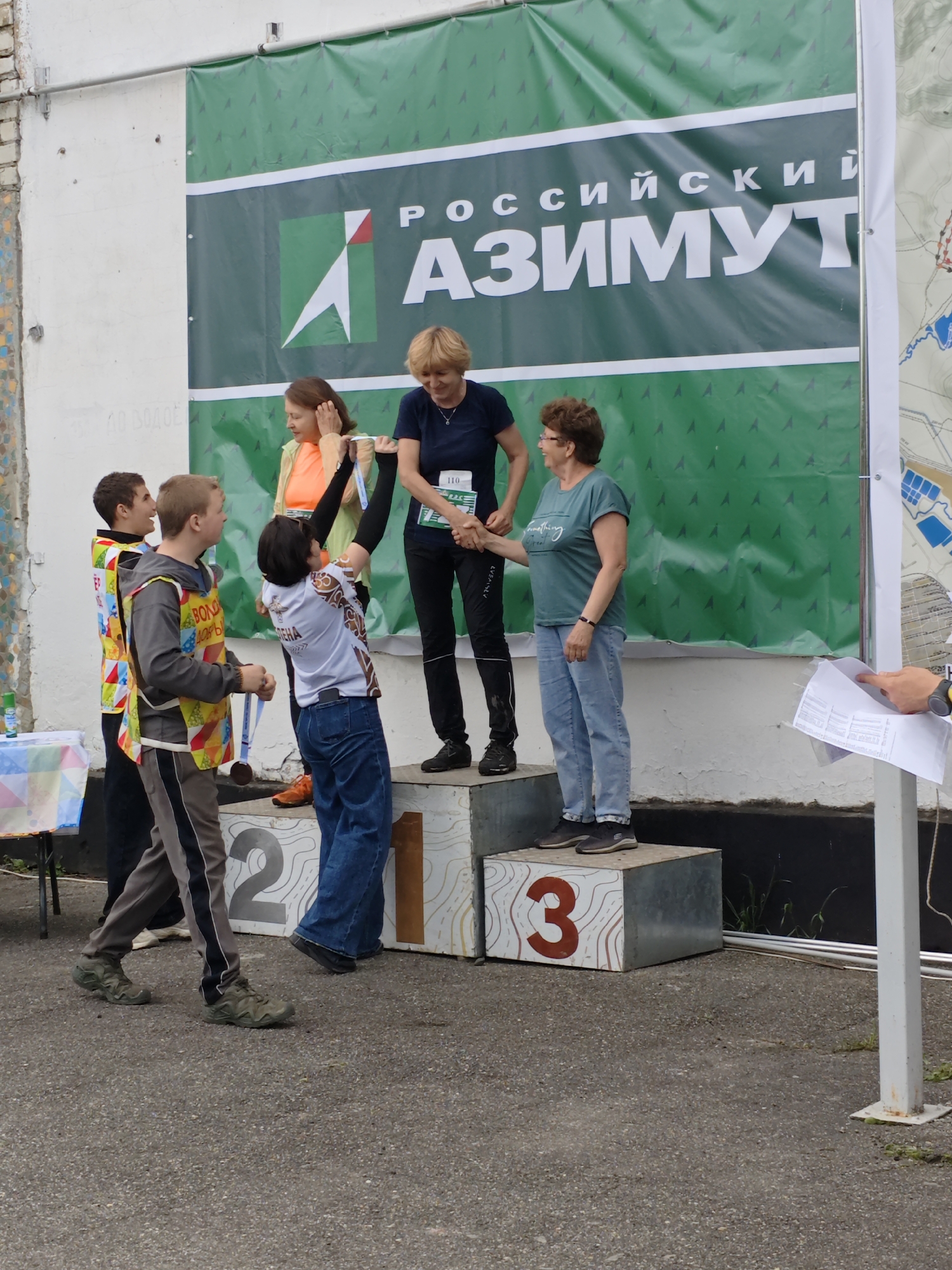 The image shows a scene in Новоекатериновская, Russia, likely an awards ceremony for a sports competition. Three people are on a tiered winner's podium labeled "1", "2", and "3". A woman in a navy top and black pants, wearing a race number 110, stands on the "1" platform, shaking hands with a woman in a teal t-shirt and blue jeans on the "3" platform. Another woman in an orange top is standing next to the "1" podium. Two young men wearing vests with colorful patterns are standing on the side of the podium. A person is handing a paper to the women standing on the "3" platform.
Behind the podium is a banner with Russian text, "РОССИЙСКИЙ АЗИМУТ", suggesting the name of the competition, with a green and white logo featuring an arrow. A map is visible on the far right side of the banner. The background includes a white brick building. A table with a blue and white tablecloth can be seen to the left of the podium. The surface appears to be asphalt.