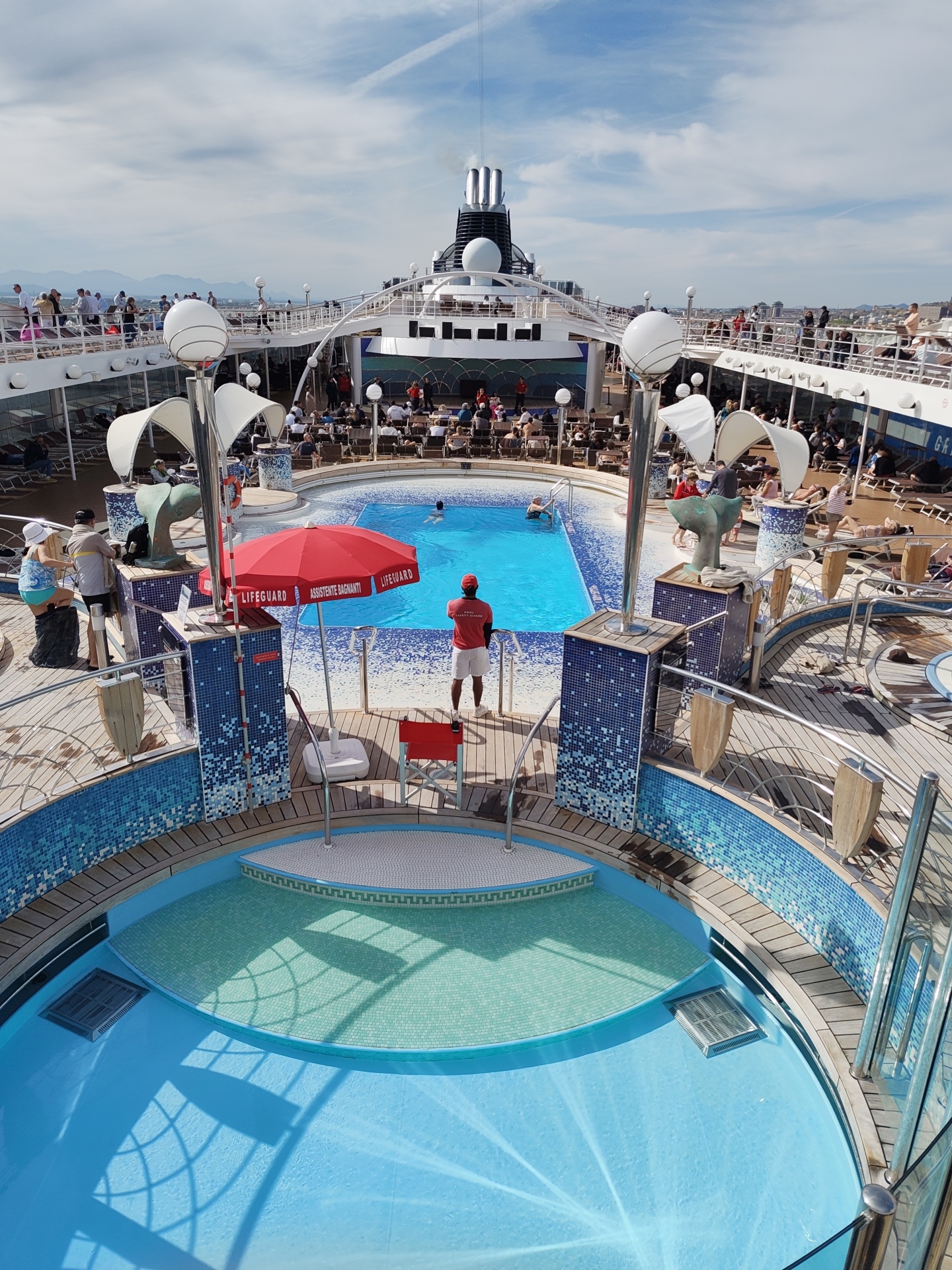 The image captures a high-angle view of a cruise ship deck with two pools, one round and one rectangular. People are swimming and lounging around the rectangular pool, where a lifeguard in a red shirt and white shorts stands watch. A red umbrella with "Lifeguard" and "Assistente Bagnanti" printed on it is nearby. The deck is crowded with people, many seated at tables on a raised platform beyond the rectangular pool. The cruise ship's chimney is visible in the background. The location is in the city of Cagliari, Italy, with distant mountains and city buildings visible on the horizon. The sky is blue with some clouds. The deck features wood flooring and blue-and-white tiled walls surrounding the pools. Some sculptures or decorative elements are also present.