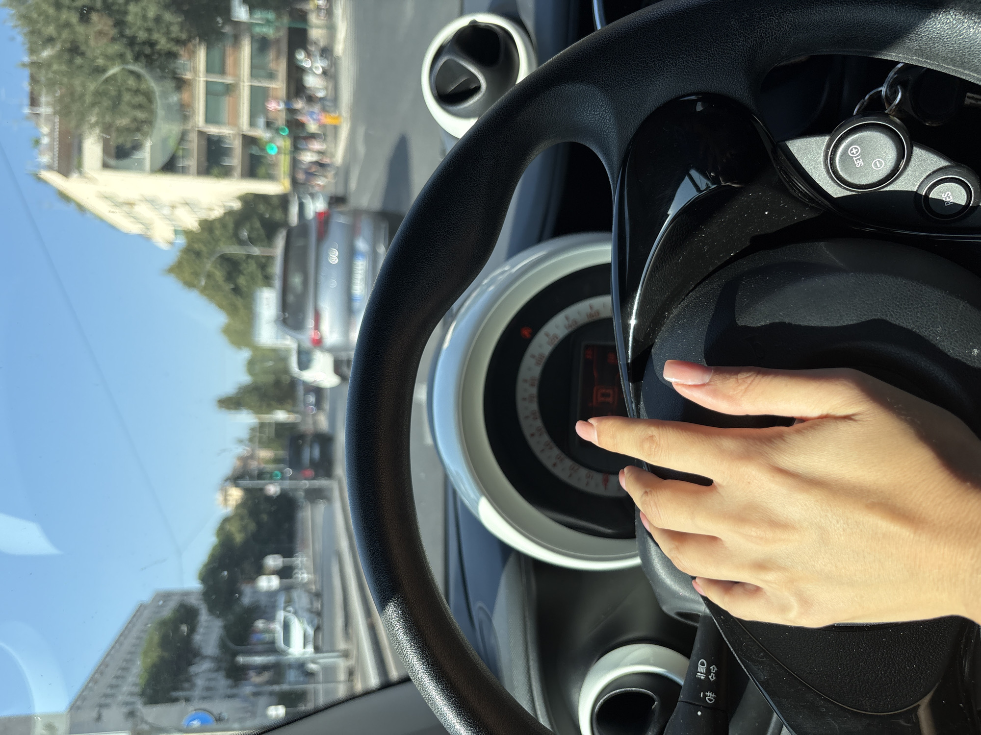 The image shows a first-person view from inside a car, with a hand resting on the steering wheel. The hand has light skin and neatly trimmed fingernails with a light pink polish. The steering wheel is black and appears to be made of textured material. To the left of the steering wheel, a part of the dashboard with controls for lights and turn signals is visible. To the right of the steering wheel, there are buttons for cruise control, marked "SET" and "RES". Behind the steering wheel, a circular instrument cluster with a digital display and a speedometer is partially visible. The car appears to be in motion on a city street under a clear blue sky. The street is lined with trees and buildings, some of which appear to be apartment buildings with balconies. The traffic includes other cars, and there are traffic lights visible in the distance. The scene is set in Rome, Italy, as indicated by the architecture and the Roman license plate on the vehicle in front.