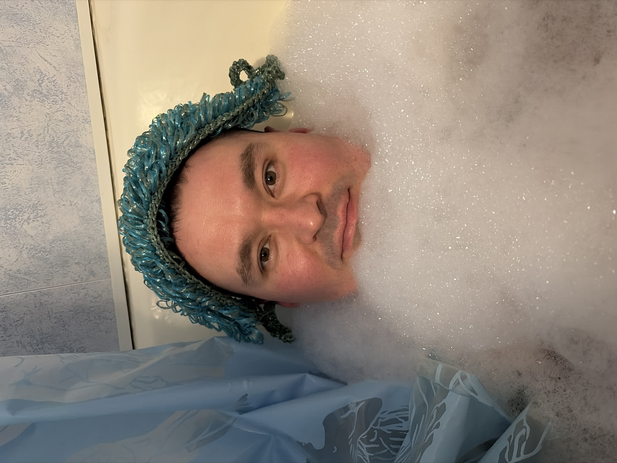 A man is lying in a bubble bath. He is wearing a blue crocheted headpiece. The setting appears to be a bathroom. There is a shower curtain with a leafy design. The location is Izhevsk, Russia.