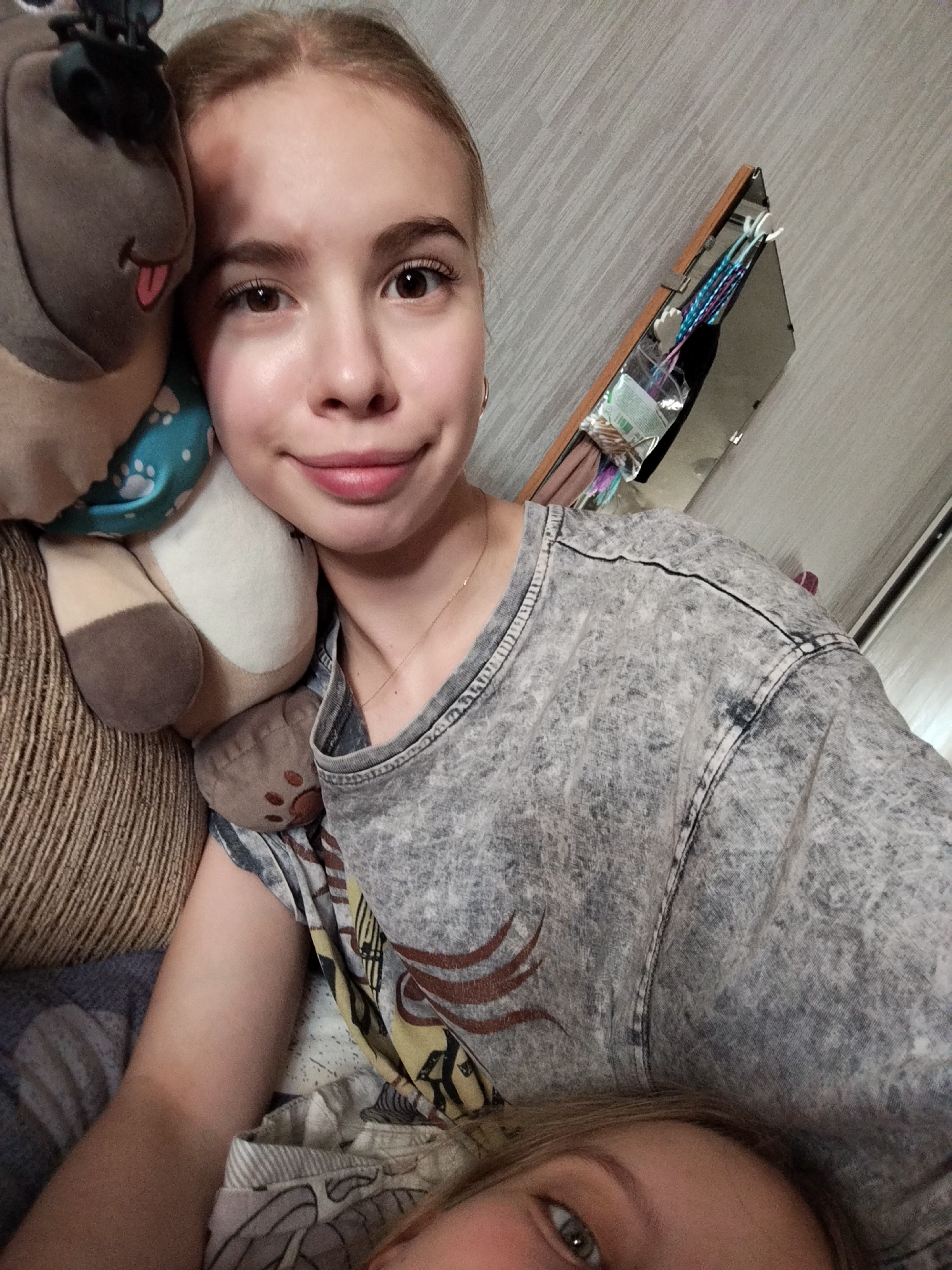A young woman is captured in a selfie, holding a stuffed animal and lying down. She is wearing a grey t-shirt with a distressed pattern. Another person's face is partially visible at the bottom of the frame, with only their eye visible. The background appears to be a room with a wooden shelf containing various items, including bags and what looks like hair accessories. The overall lighting suggests an indoor setting.