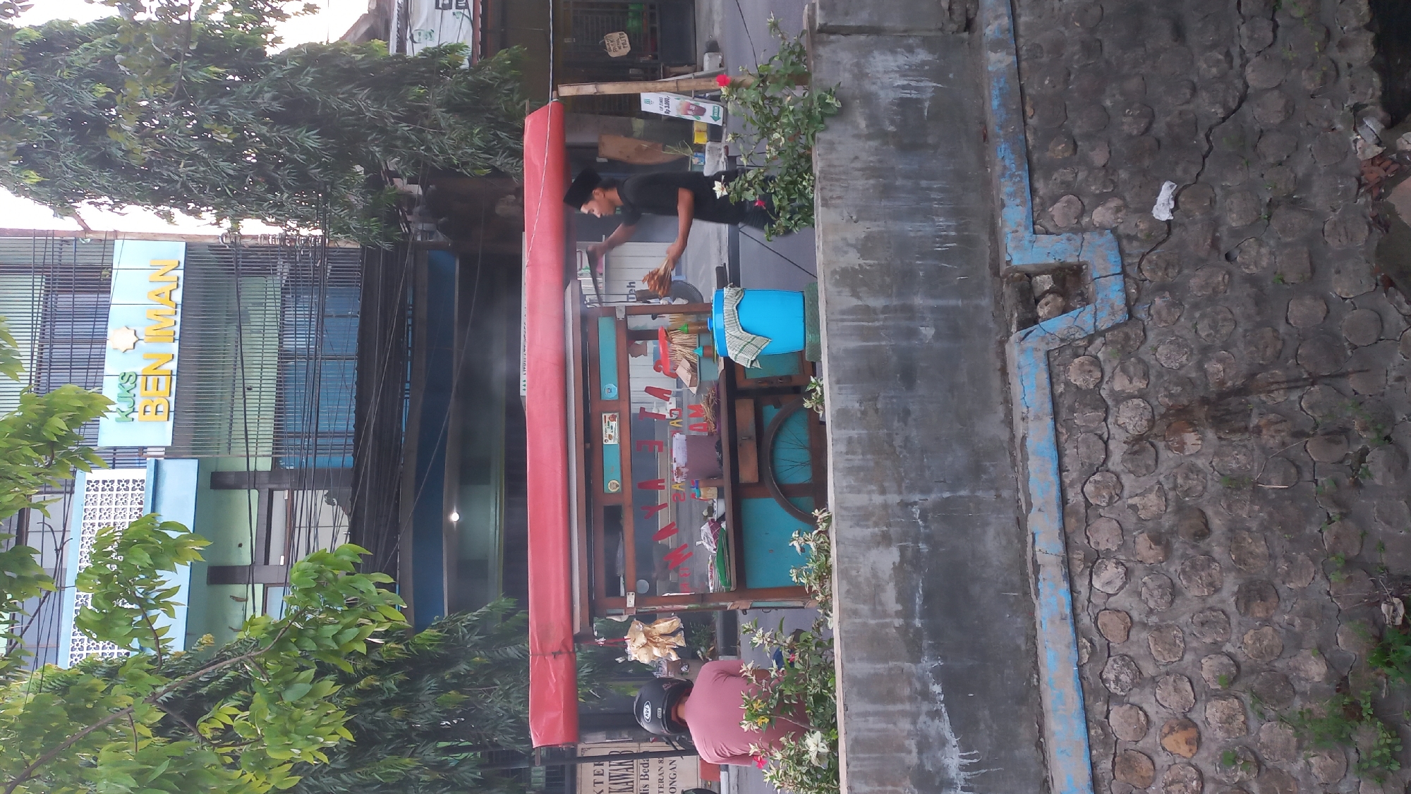 In Jetis Indah, Indonesia, a street food cart is depicted beside a busy road. The cart has a red canopy and a turquoise base. A vendor, wearing a black shirt and peci, is standing and serving food. In front of the cart, a person wearing a pink shirt and a black helmet is seated on a scooter. The scene also features a stone wall in the foreground, with foliage growing around it. A building with a sign that reads "KUKS BEN IMAN" is visible in the background. The overall impression is of a vibrant street scene in an Indonesian city.