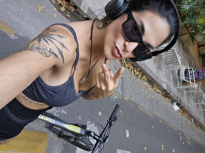 A woman wearing dark sunglasses and headphones poses with a bicycle. She's wearing a gray sports bra and black shorts. A leaf tattoo is visible on her upper left arm. She is holding up two fingers in a peace sign. The background features a street with scattered leaves and metal chairs near a sign.