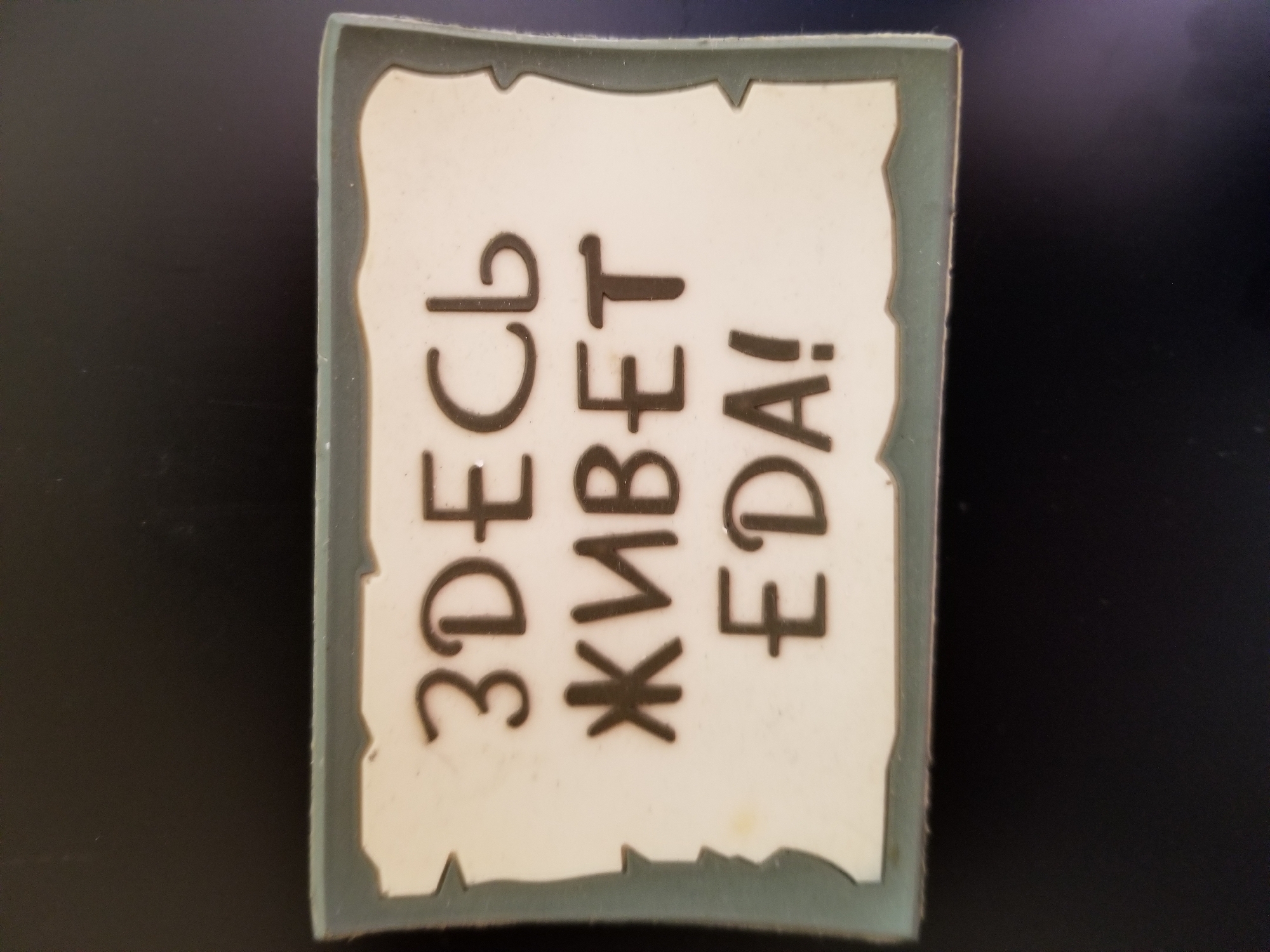 A sign with Russian text is displayed against a dark background. The sign is rectangular with a decorative border. The text on the sign reads "ЗДЕСЬ ЖИВЕТ EDA!" which translates to "EDA LIVES HERE!". The letters are dark brown and are capitalized. The sign appears to be a novelty item or a humorous decoration. There are no people or other objects in the image. The location in Tiraspol, Moldova, is not depicted in the image.