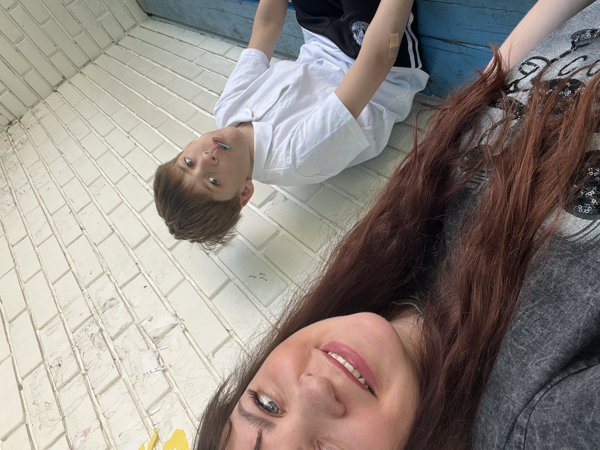A selfie depicts a woman with long brown hair and a boy in an outdoor setting. The woman is wearing a gray shirt and smiling at the camera. The boy is positioned above her in the frame, wearing a white shirt, black pants with white stripes, and a bandage on his arm. He has a green object in his mouth. They appear to be against a white brick wall. Above them is a blue painted wooden structure. The location is Noviy, Russia.
