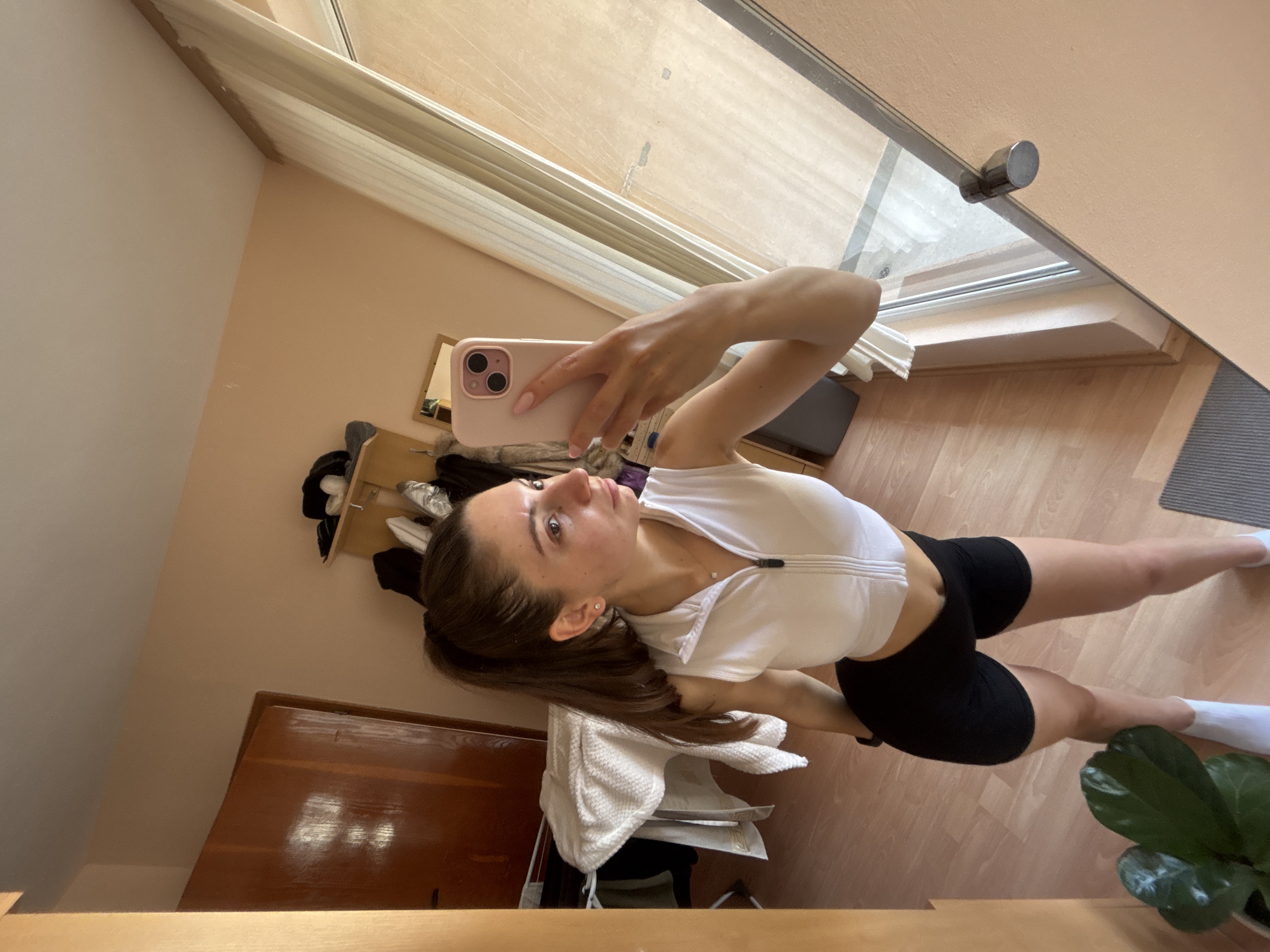 A young adult female, with dark brown hair tied in a high ponytail, stands in a light peach-walled room in Varna, Bulgaria, taking a mirror selfie. She is captured from the mid-thigh up, slightly angled, wearing a white cropped sleeveless top with a half-zip closure, black athletic shorts, and white ankle socks. A small, square-shaped pendant necklace and a silver stud earring are visible. Her right hand holds a pink smartphone, pointed towards the mirror, with her left hand resting on her left hip. Her gaze is directed to her right, away from the phone.
The room features light brown laminate flooring and a large rectangular mirror reflecting the scene. To the left of the subject in the reflection is a dark brown wooden door, partially obscured by a white towel draped over a clothes drying rack with other dark garments. A prominent light wooden door frame borders the left side of the mirror. In the reflected background to the right, a large window with light-colored blinds or shades is visible, offering a glimpse of an indistinct exterior. A small, framed mirror is reflected on the wall above the window. A cluttered shelf unit is visible in the upper right corner of the reflection. In the foreground, outside the mirror's reflection, a potted plant with large green leaves sits in a white pot on the floor, and a dark gray mat is partially visible in the lower right.