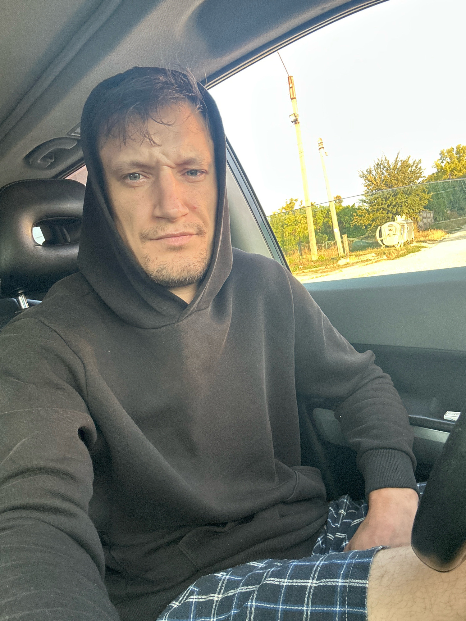 A selfie taken from inside a vehicle shows a man sitting in the driver's seat. He is wearing a dark hooded sweatshirt and blue and white checkered shorts. The man has light skin, light-colored eyes, and short, dark hair that appears slightly disheveled. His facial expression is one of mild disgruntlement. Through the car window, a rural or semi-rural landscape is visible, with trees, a fence, and utility poles under a bright, clear sky. One of the poles has what appears to be a street light or camera fixture attached. A white cylindrical object, possibly a trash bin or storage container, is also visible in the background. The interior of the car is dimly lit, with the headrest of the passenger seat visible to the left of the man. The ambient light suggests it is daytime, likely late afternoon or early morning due to the angle of the sun.