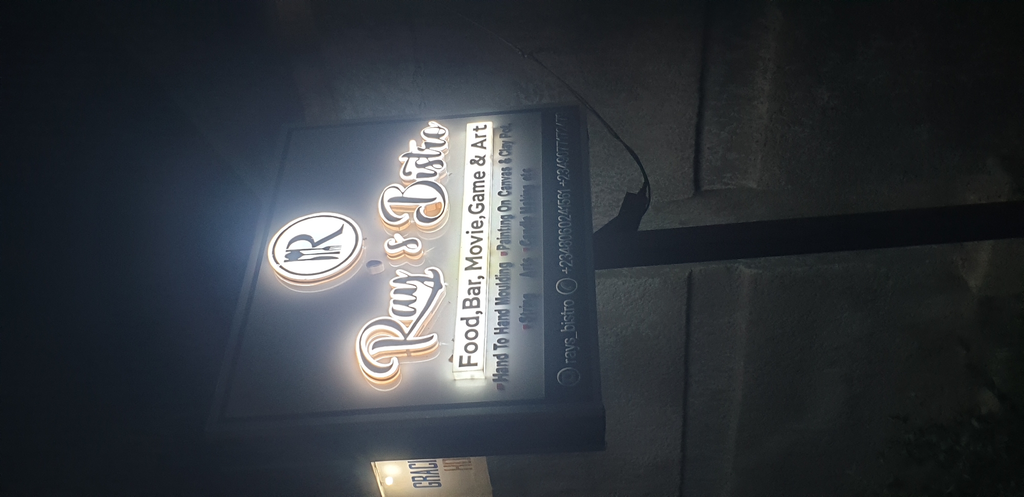 The image depicts a nighttime scene in Narayi, Nigeria, featuring a brightly lit sign for "Ray's Bistro" mounted on a pole against a dark, textured wall. The sign displays "Ray's Bistro" in prominent, illuminated script with a circular logo above it that contains an "R" and what appears to be a fork and knife. Below the name, the sign lists services: "Food, Bar, Movie, Game & Art". Further details for "Art" services are visible: "Hand To Hand Moulding," "Painting On Canvas & Clay Pot," "String Arts," and "Candle Making, etc." At the bottom of the sign, an Instagram handle "rays_bistro" is present, along with two phone numbers: "+2348060241561" and "+234907777247". To the left of the main sign, a partial view of another lit sign is visible, displaying "GRACI" and "HO," suggesting "Grace Hotel" or similar establishment. The immediate foreground and background are dark, with minimal discernible features beyond the illuminated signs and the textured wall. No individuals are visible in the scene.