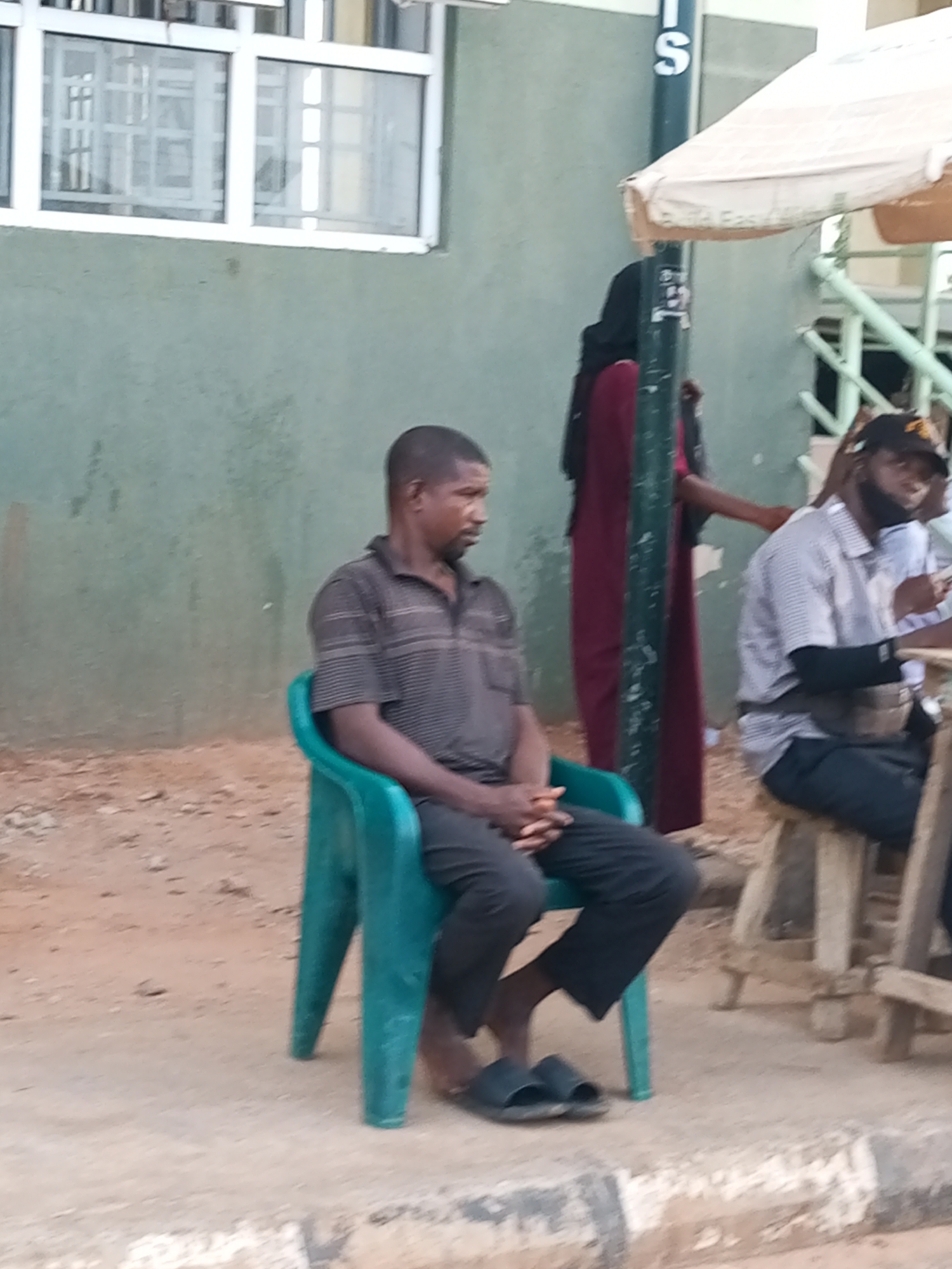 In Gombe, Nigeria, a man sits on a turquoise plastic chair. He is wearing a dark, horizontally striped short-sleeved shirt and black trousers. His feet are clad in black slides. Behind him, a woman in a dark hijab and a maroon garment stands near a lamppost. To his right, another man, wearing a checkered shirt, a black cap, and a dark face mask, is seated on a wooden stool. The background features a green-painted wall with barred windows, suggesting a building. The ground is a mixture of dirt and concrete, with a curb visible in the foreground. An umbrella provides shade in the upper right portion of the image.