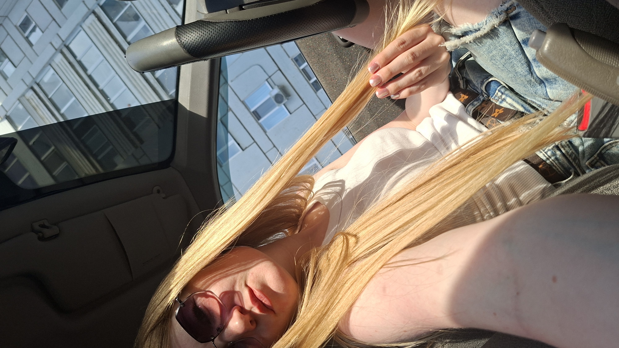 A light-skinned person with long, blonde hair and sunglasses is sitting in the passenger seat of a vehicle. The person is holding a strand of their hair. The interior of the vehicle is visible, including the sun visor, handle, and seat. Outside the vehicle window, a tall building with multiple windows is seen. The photo was likely taken during the day due to the bright sunlight. It is not possible to verify that the location is in Chaikovsky, Russia from the image alone.