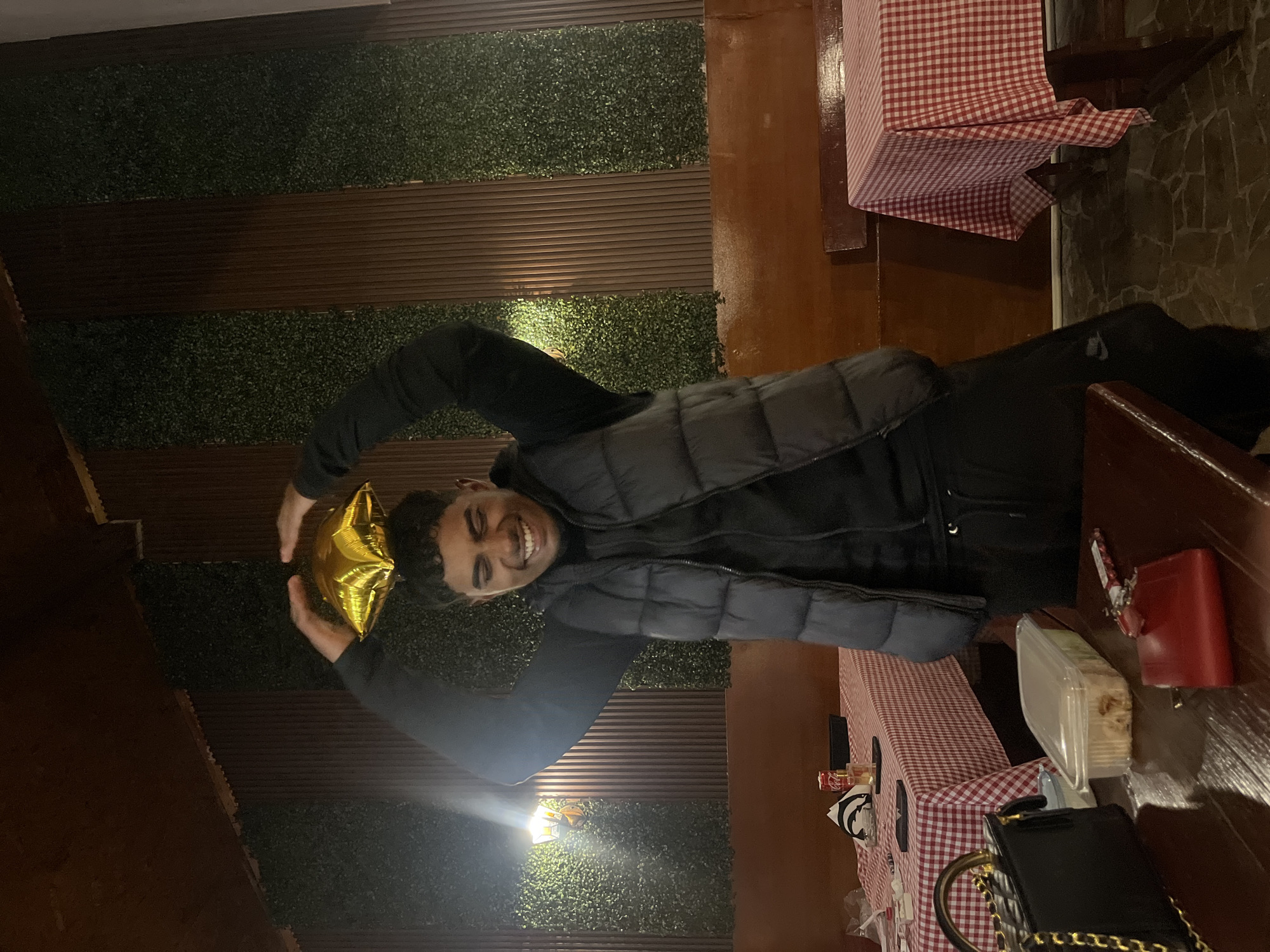 The image shows a man indoors, smiling and holding a gold star-shaped balloon. He is wearing a dark jacket and vest. He is seated at a table covered with a red and white checkered tablecloth. On the table are various objects including a black purse with a gold chain, a clear plastic container, and a red box. The wall behind the man is decorated with panels of alternating brown wood and artificial green foliage. The location is potentially a restaurant or dining establishment within Lozarevo, Bulgaria.