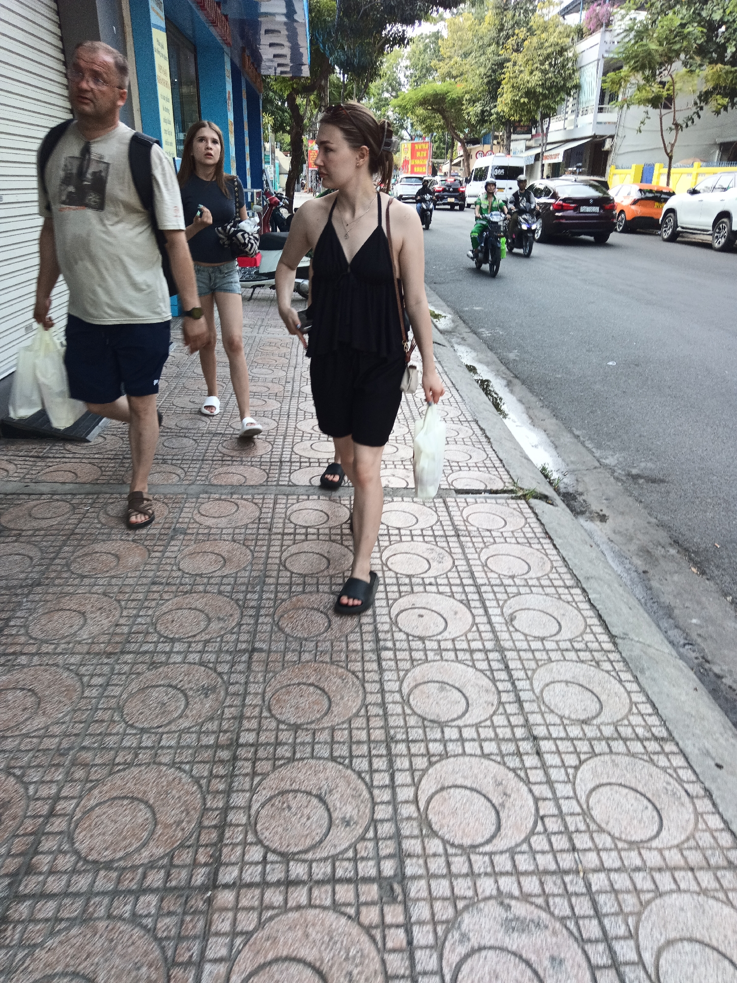 The image depicts a street scene in Nha Trang, Vietnam. Three individuals are walking on a patterned sidewalk adjacent to a road with vehicle traffic.
In the foreground, a man with glasses and a backpack is walking to the left, holding a white plastic bag. He is wearing a light-colored t-shirt and dark shorts. To his right, a young woman with long brown hair is walking, looking to her right. She is wearing a dark top and denim shorts. Further to the right, another woman with her hair pulled back is walking away from the camera, holding a white plastic bag and wearing black shorts and a black top.
The sidewalk is paved with square tiles, each featuring a circular pattern. The road beside the sidewalk shows various vehicles, including motorcycles and cars. The background includes buildings with blue accents, trees, and a cloudy sky. The overall impression is that of a typical urban street environment in Vietnam.