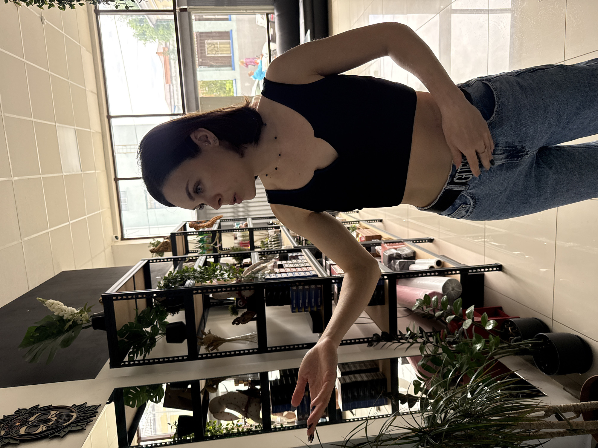 A medium shot depicts a woman indoors, in what appears to be a retail space or store. She is standing, leaning slightly forward, and looking down. The woman is wearing a black cropped tank top and blue jeans with a black belt that has the word "GUESS" visible. Her hair is dark, and she has an earring visible in one ear.
She is reaching towards a shelf unit filled with various items, including plants, decorative objects, and what appear to be rolls of fabric or paper. The setting includes a tiled floor, a window that looks out onto a street scene in what appears to be an urban setting. Based on available information the city could be Rostov-on-Don, Russia.