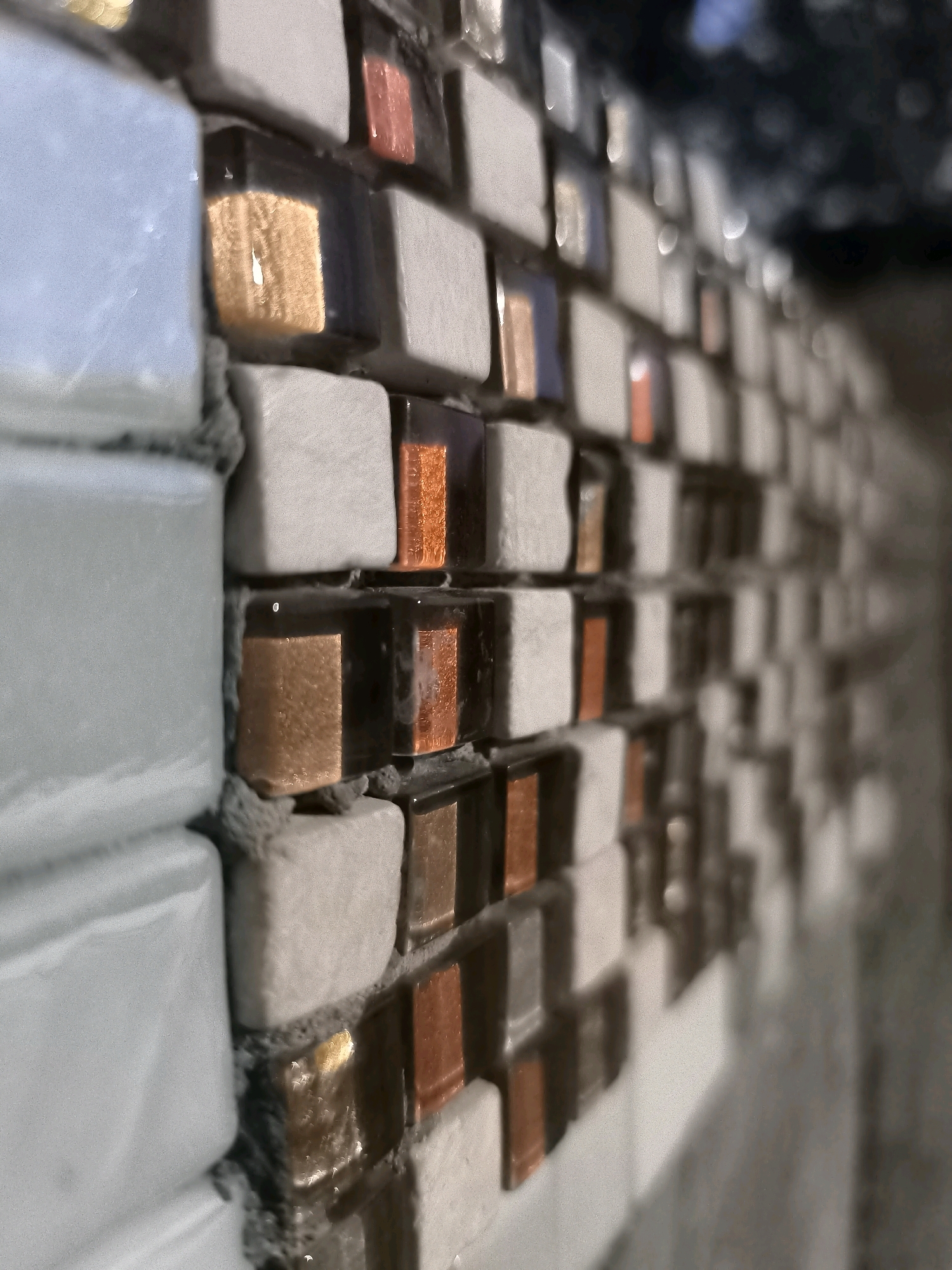 The image shows a close-up, side-view of a mosaic wall. The mosaic consists of small, square tiles of various colors and finishes, including light blue, grey, white, copper, and black. The tiles are arranged in a staggered pattern, with the grout between them visible. The perspective is from the left side of the wall, looking towards the right and slightly upwards. The lighting highlights the glossy surfaces of some of the copper and black tiles, creating reflections. The background is blurred and out of focus, suggesting a shallow depth of field. The location in Namdanak, Uzbekistan, is not discernible from the visual information presented in the image.