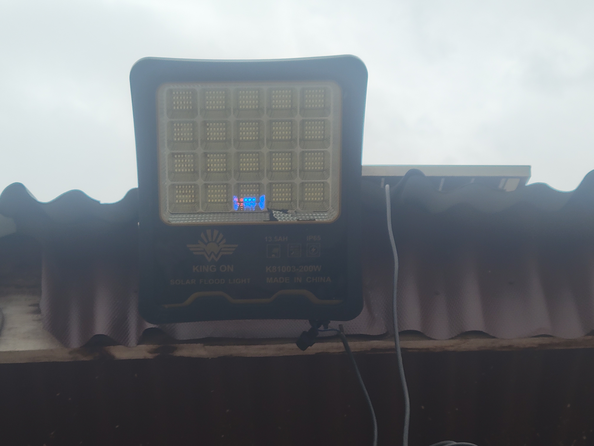 The image shows a solar floodlight mounted on a corrugated roof. The floodlight is black and rectangular with a grid of LEDs on its front. The brand name "KING ON" is visible, along with the text "SOLAR FLOOD LIGHT", "13.5AH", "IP65", "K81003-200W", and "MADE IN CHINA". The sky in the background is overcast, suggesting cloudy weather. There are no people visible in the image. The setting appears to be an outdoor area, possibly a residential or commercial building, given the rooftop installation. The floodlight is the primary object of focus. The time of day is unclear due to the overcast sky.