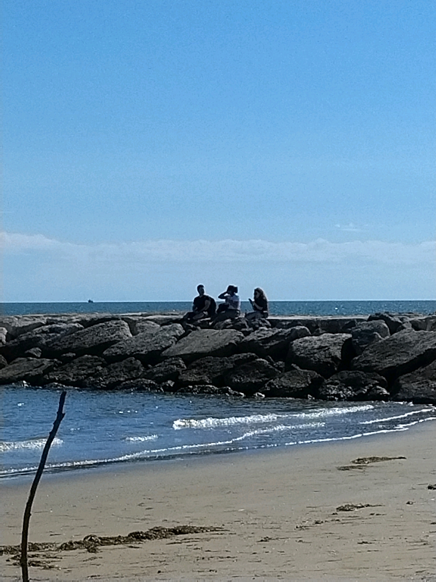 The image depicts a breakwater made of large, dark rocks extending into the sea. Three individuals are seated on top of the breakwater, facing the water. The person on the far left is wearing dark clothing, the middle person is wearing a white top and dark bottoms, and the person on the right is wearing dark clothing. The sea is calm with small waves breaking near the shore. The shore is sandy, with some seaweed visible. A thin, dark stick is propped upright in the sand in the foreground. In the distance, on the sea, a small boat is visible. The sky is clear and blue with a few wispy clouds. This scene is consistent with the breakwaters found along the coast of Caorle, Italy, which are often used as seating areas by visitors and locals.