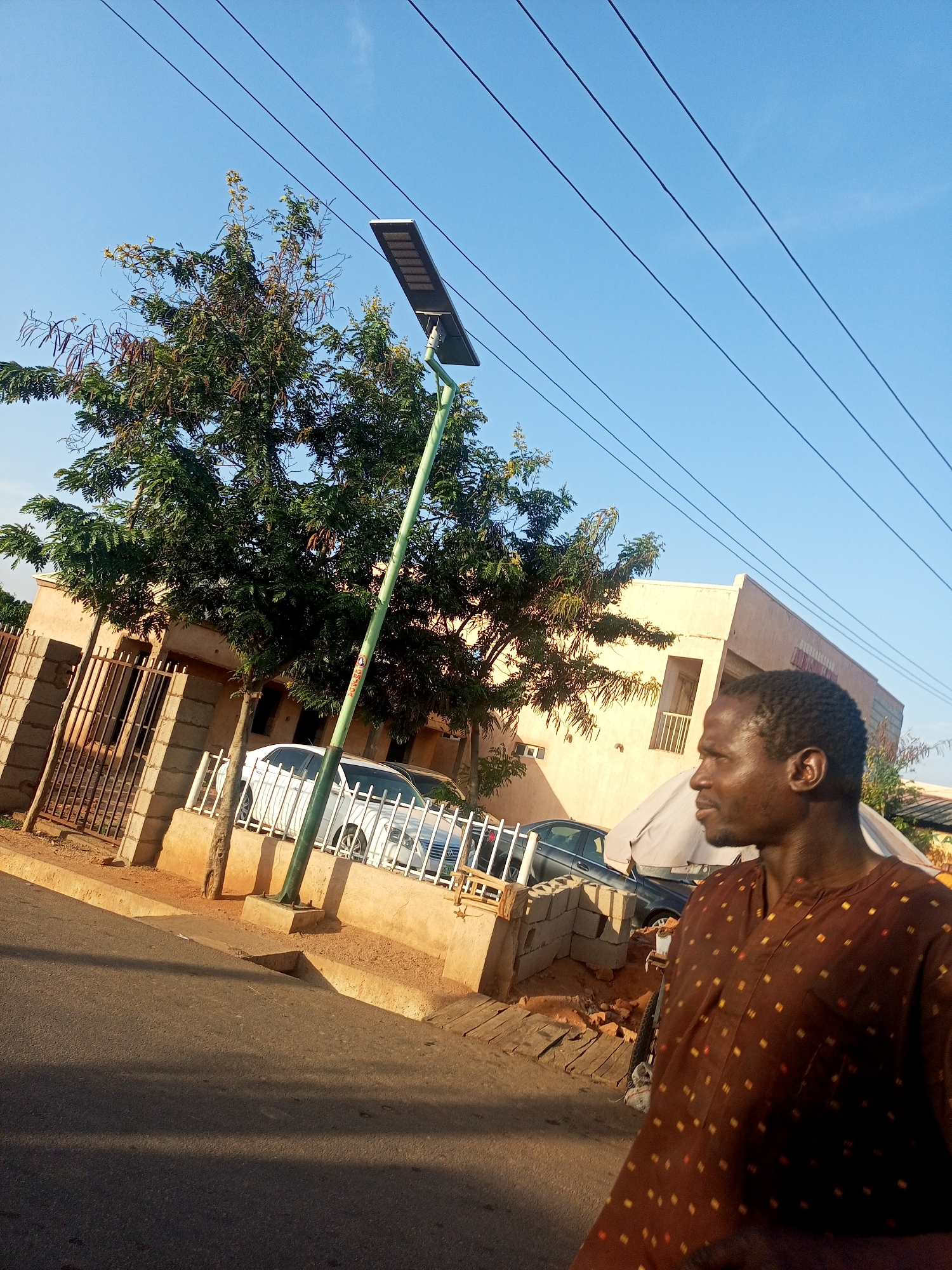 A street scene in Gombe, Nigeria, is captured during what appears to be the late afternoon, given the warm, directional light and prominent shadows. In the foreground on the right, a dark-skinned man is seen in profile from the chest up, looking towards the right. He wears a loose-fitting brown shirt adorned with small, lighter-colored rectangular patterns.
Behind the man and extending towards the left is a green street light pole, equipped with a solar panel and an LED lamp fixture. Above, several power lines stretch across a clear, bright blue sky. A large tree with dense green foliage and visible dried seed pods stands in the midground, partially obscuring a light-colored building that features multiple windows and a balcony railing on an upper floor.
The property in front of the building is secured by a fence made of concrete blocks and vertical metal bars. Beyond this, a white car and a darker vehicle are parked, separated from the street by another white metal fence. The immediate foreground shows an asphalt road, partially covered in shadows, leading to a dirt shoulder containing concrete debris and the base of the street light pole. No clearly legible text is visible on any objects.