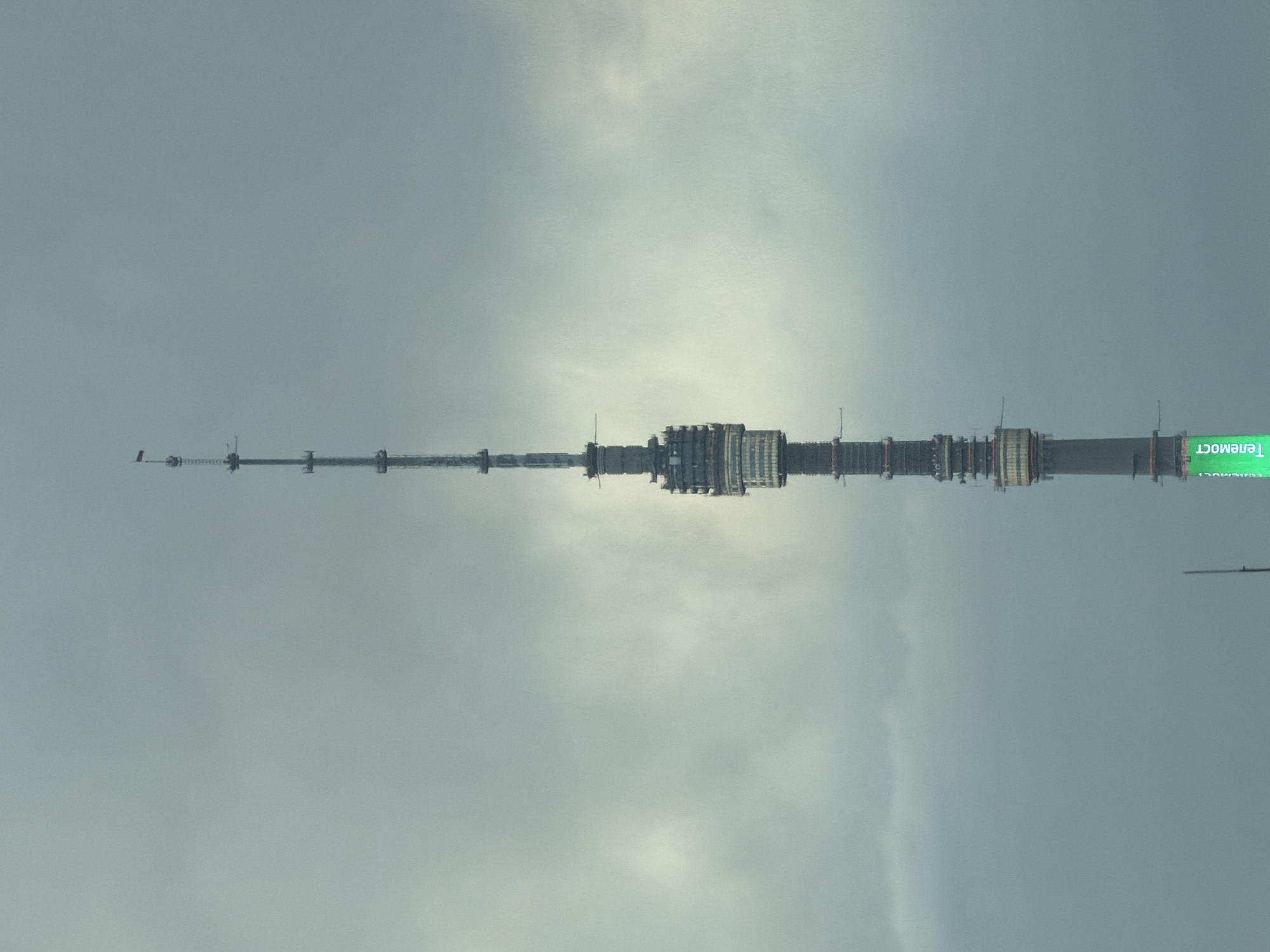 A low-angle shot depicts the Ostankino Tower against a gray, overcast sky. The tower extends horizontally across the frame with its slender, lattice-like mast tapering to the left and the wider base of the tower on the right. A green, horizontally oriented sign with the word "Телемост" (Telemos) is visible. No people are visible.