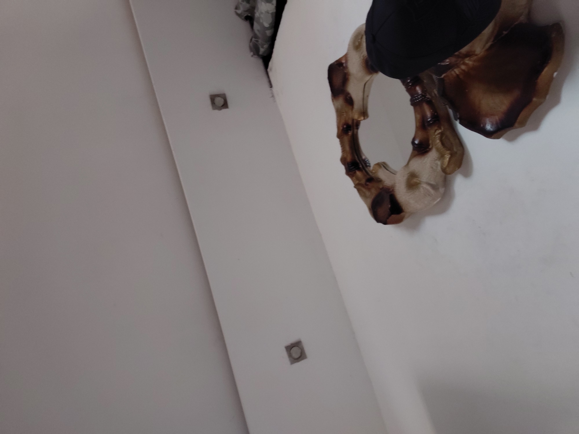 The image displays an indoor scene, likely a room corner, viewed from a rotated perspective. The dominant colors are white, brown, beige, and black.
The primary subject is a distinctive, organically shaped decorative object, possibly a mirror or a wall sculpture, mounted on a white wall on the right side of the frame. This object has an irregular, somewhat circular form, resembling carved wood or a large natural fungus. It features contrasting textures and colors: glossy, deep brown sections are interspersed with lighter, matte beige or cream-colored areas, suggesting a natural material with a polished finish. A portion of a black item, perhaps a hat or fabric, is seen resting on the top edge of this decorative piece.
On the left side of the image, another white surface, possibly a wall or ceiling, forms an angle with the first. Embedded within this surface are two small, square fixtures, which appear to be recessed lights or vents, positioned vertically apart. In the upper-left corner, a sliver of dark fabric with a subtle pattern, possibly camouflage, is visible.
The scene is static, with no activity depicted. The lighting is soft and ambient, characteristic of an interior setting, and there are no specific visual cues to indicate the time of day or weather. No visible text is present in the image. The location context for this media file is Santa Ana, Venezuela.