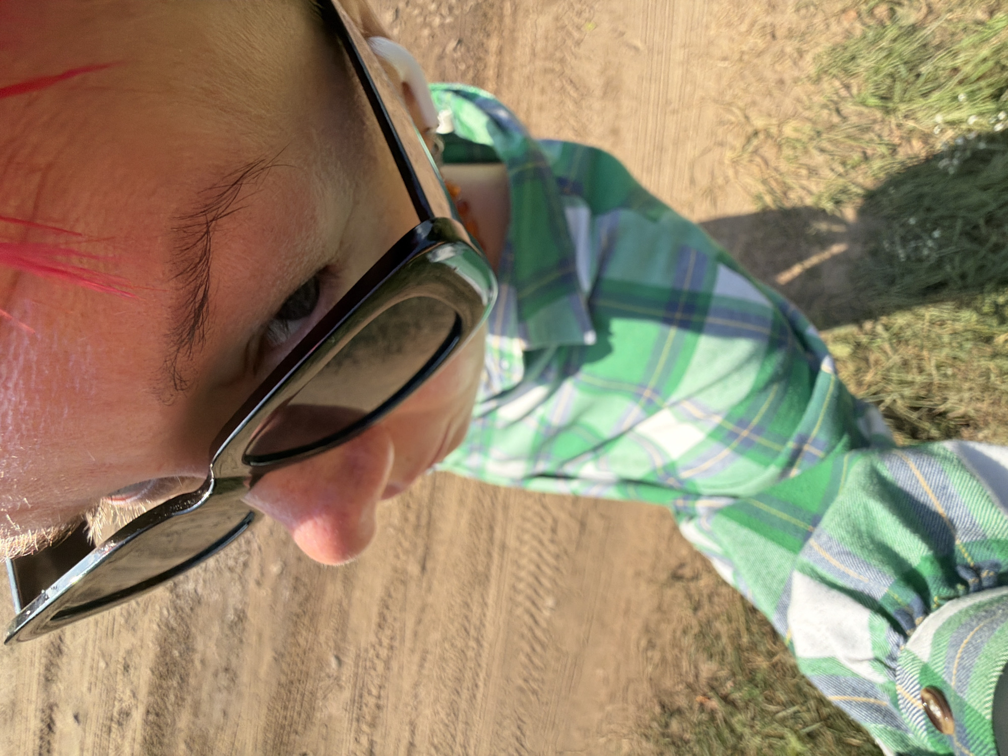 A person, viewed from a high angle, is wearing black sunglasses and a green and white plaid shirt. They have bright pink hair. The person is outdoors, standing on a dirt path with some grass on the side. The scene appears to be the city of Chaikovsky, Russia, based on the context provided. The lighting suggests it is daytime.