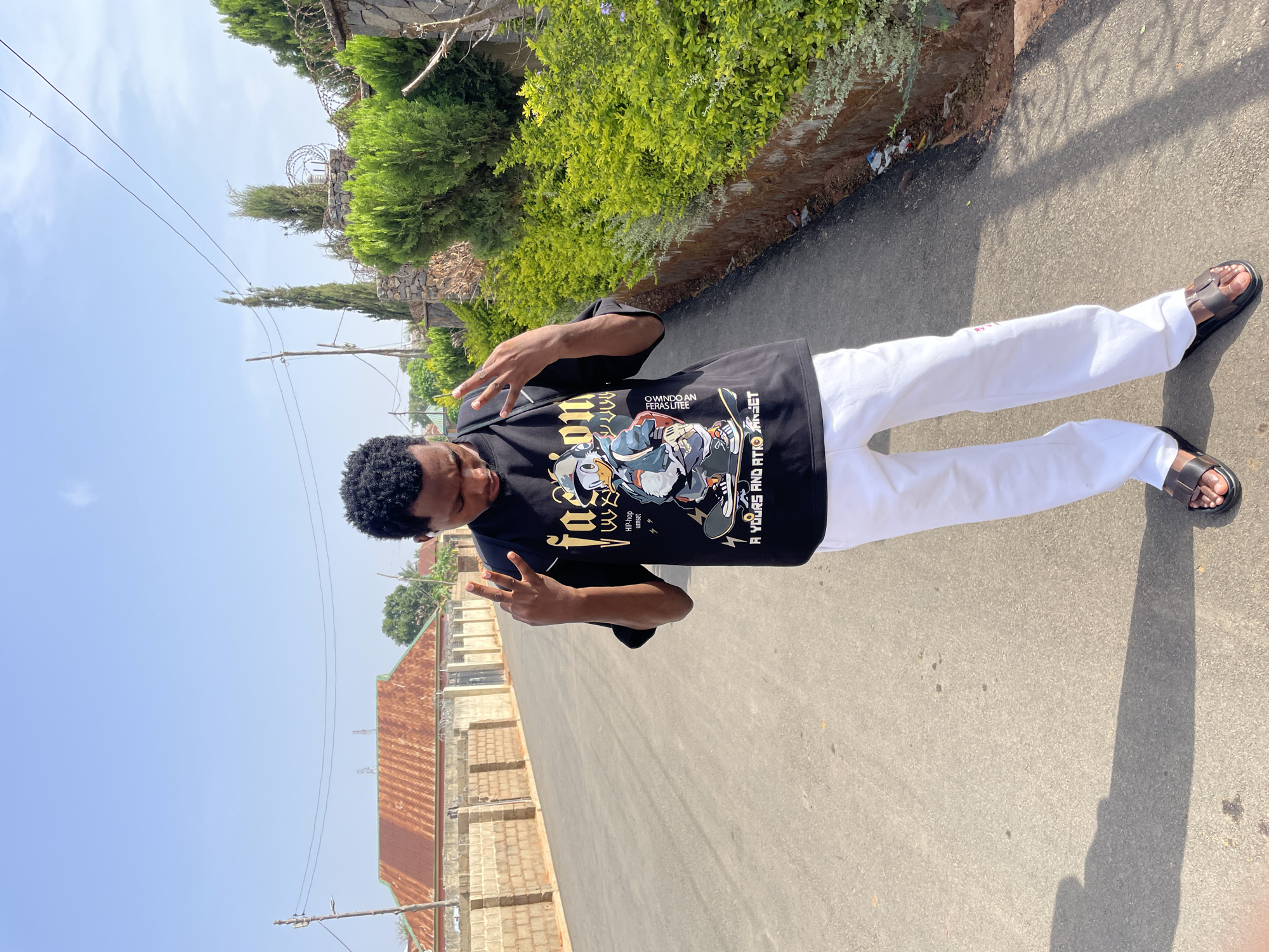 The image shows a young man posing on an asphalt road under a bright blue sky, likely in Jos, Nigeria. The photo appears rotated 90 degrees clockwise. The man, with short, dark, curly hair, stands facing the camera, making "peace" or "victory" signs with both hands. He is dressed in a black t-shirt featuring colorful, cartoon-style graphics, white loose-fitting pants, and dark sandals.
The outdoor setting is bathed in bright sunlight, with long shadows stretching across the asphalt, indicating it is likely midday or early afternoon. The road is bordered by green foliage on both sides. To the right, a stone or concrete block wall topped with barbed wire is visible behind a dense green hedge. On the left, older buildings with reddish-brown corrugated iron roofs and brick walls are partially seen. Overhead, power lines traverse the clear blue sky, supported by a wooden utility pole.
Text visible on the black t-shirt includes "facson", "HIP-hop", "urnset", "OWINDO AN", "FERAS LITEE", and "A YOURS AND ATKOGET", suggesting brand names or thematic elements. The man's posture and gestures convey a relaxed and confident demeanor, posing for the photograph in what appears to be a casual, everyday street scene in Jos.