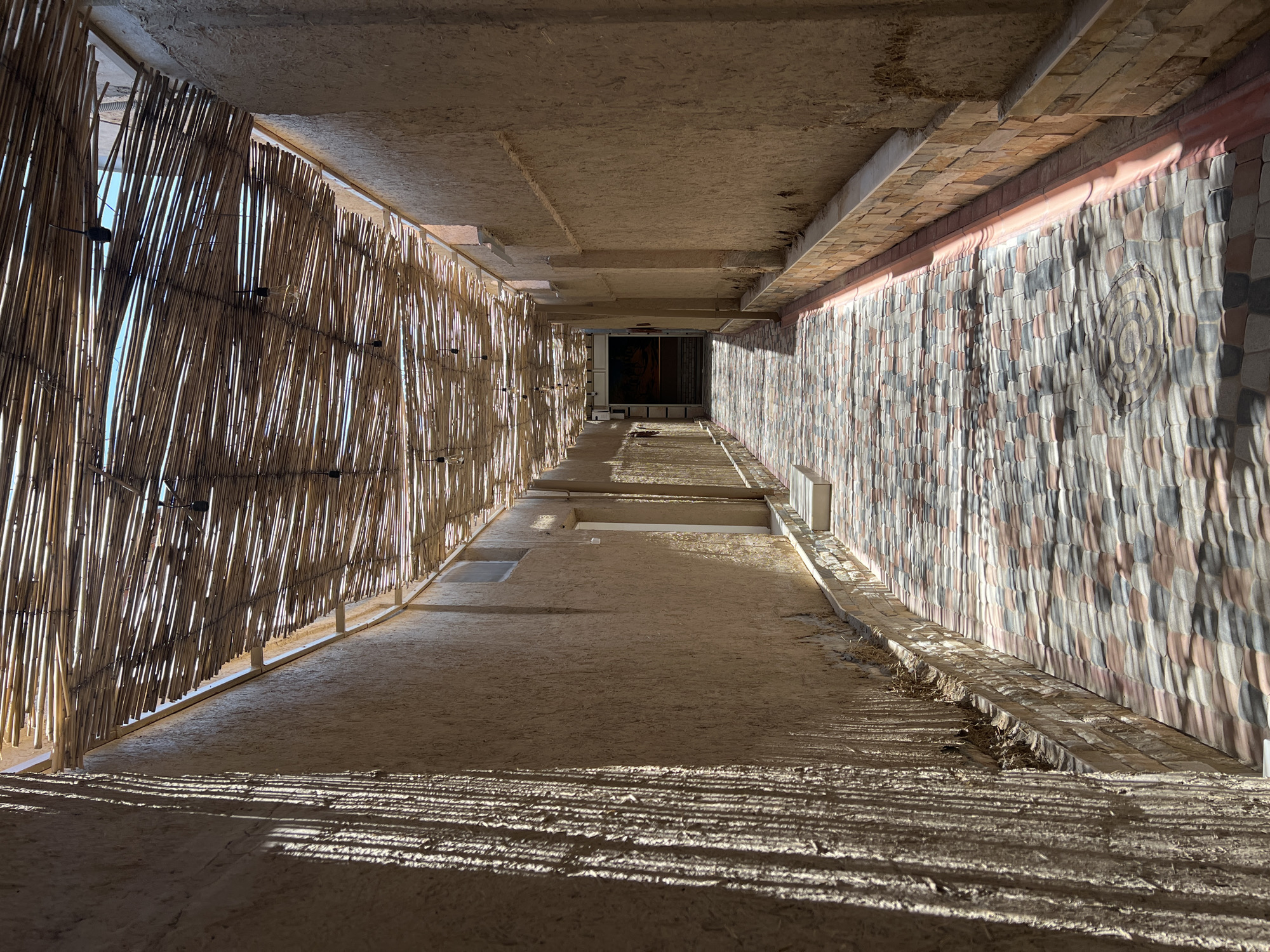This media file captures a downward perspective of a sun-drenched corridor, likely part of a building under construction or with an unfinished aesthetic. The scene is characterized by strong contrasts between light and shadow, with parallel lines of light filtering through a bamboo-like screen on the left, casting distinct striped patterns on the sandy ground. This screen is composed of numerous vertical, thin wooden elements, with visible light bulbs strung across it, suggesting an outdoor or semi-outdoor setting.
The right wall of the corridor is adorned with a mosaic of irregularly shaped, textured tiles in varying shades of grey, pink, and beige, creating a visually dynamic surface. Above, the ceiling is a rough, concrete structure with visible beams and formwork, further indicating an unfinished state. The floor is sandy and uneven, with some debris and dry vegetation visible.
The play of light and shadow suggests a time of day when the sun is relatively low in the sky, possibly late afternoon or early morning. The bright sunlight and lack of visible precipitation point to clear weather. The architectural style, with the bamboo screen and mosaic tiling, alongside the sandy ground, hints at a location in a warm climate, consistent with the provided location context of Samarkand, Uzbekistan. There is no visible text in the image. The overall impression is one of raw, natural materials and the interplay of light within an architectural space.