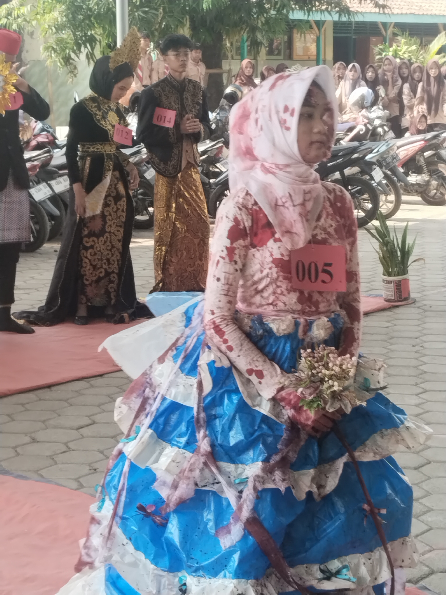 In Pakulaut, Indonesia, a person wearing a white hijab and a garment stained with red paint, possibly representing blood, stands in the foreground. The garment is layered with blue and white plastic, resembling a skirt. A small bouquet of flowers is held in their hand, and a red placard with the number "005" is pinned to their chest. Behind them, other individuals are present, some wearing traditional Indonesian attire, including a person in a black and gold outfit with a golden headdress and another in a black jacket with gold embroidery and a golden sarong. Red placards with numbers "012" and "014" are visible on these individuals. Motorcycles are parked in the background. The scene appears to be an outdoor event, possibly a parade or competition, taking place on a paved area with a red carpet and potted plants visible. The overall lighting suggests it is daytime.