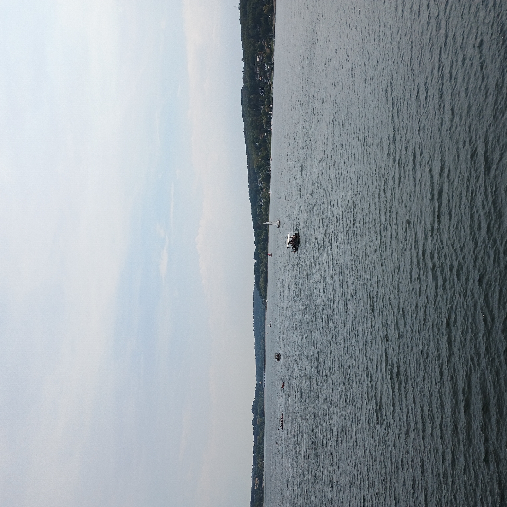 A wide shot captures a serene lake, likely Möhnesee in Germany, on a cloudy day. The water, with subtle ripples, stretches towards a distant, tree-lined shore dotted with buildings. Several boats are scattered across the expanse, including a small, open-air vessel with people aboard in the middle of the lake. To the left, a larger boat appears to be carrying a group of individuals, possibly a rowing crew. In the distance, a few sailboats are visible. The sky is overcast with soft clouds, creating a diffused light across the scene.
