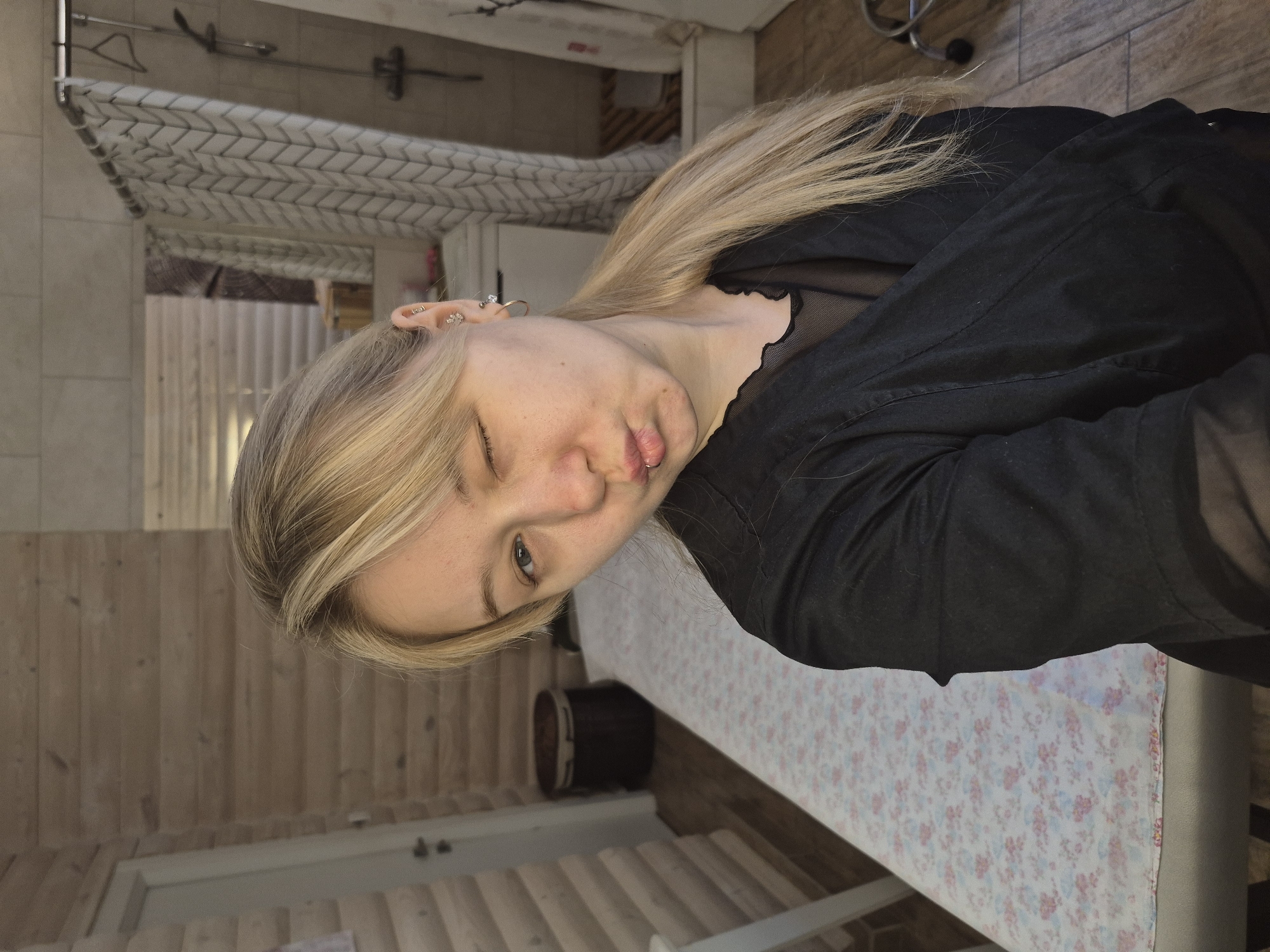 A blond-haired woman in a black shirt winks at the camera. She is likely indoors in a room with wood paneling, a shower with a patterned curtain, and a patterned tablecloth on a table or surface in front of her. It is not possible to confirm the location in Chaikovsky, Russia from the image.