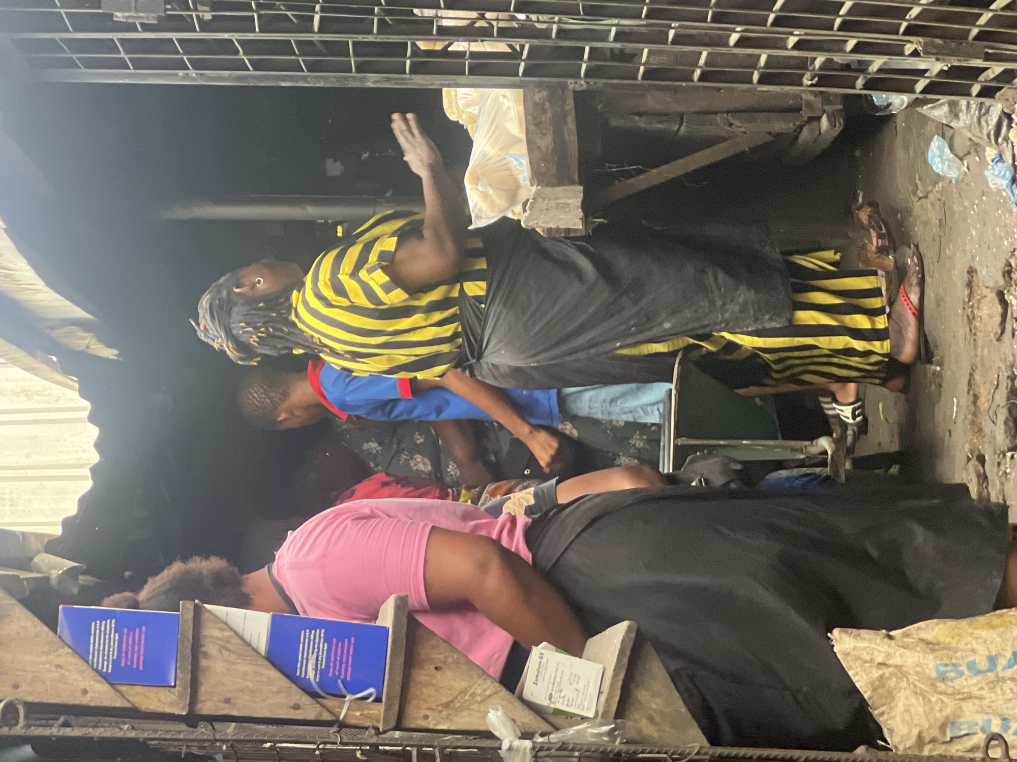 In Nkpor, Nigeria, a marketplace scene unfolds. A woman in a bright pink t-shirt and a dark skirt stands facing away from the camera. To her left, on a wooden shelf, are two blue boxes, likely containing medication, and a smaller white box labeled "Zumatem-DS". Behind her, several other individuals are visible, including a woman in a black and yellow striped outfit with a black apron, who appears to be interacting with someone to her right, gesturing with her hand. The environment is dimly lit, suggesting an indoor or covered market space, with corrugated metal roofing visible overhead and a metal gate on the right side of the frame. The ground is uneven and appears to be a dirt or concrete surface. Several plastic bags and other debris are scattered on the ground.