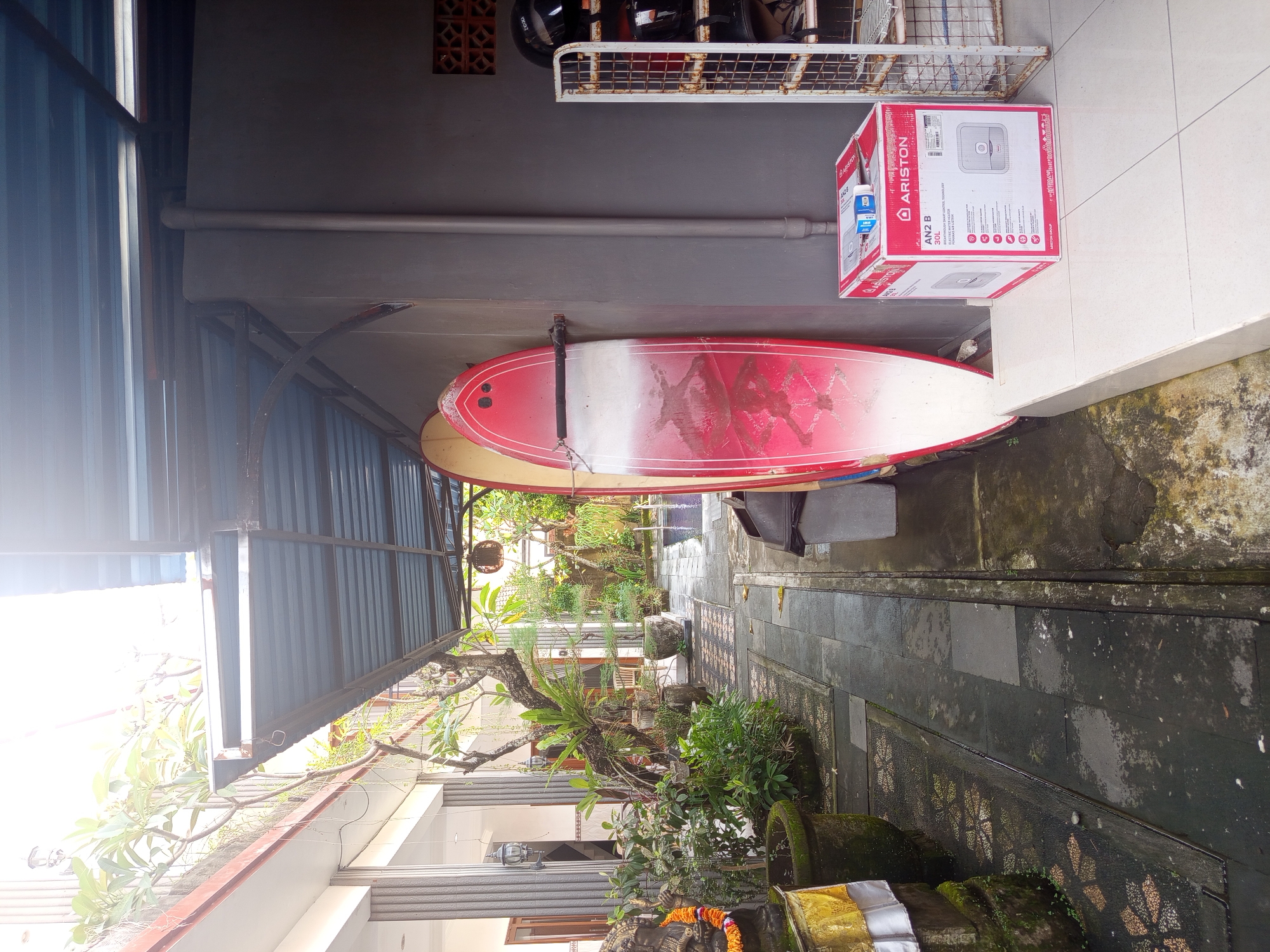 The image shows a narrow alleyway in North Kuta, Indonesia. On the left is a building with a corrugated metal roof and visible plants. The alley floor is paved with dark, textured tiles. On the right, a tiled wall shows signs of moisture and weathering. A surfboard and a boxed water heater (Ariston AN2 B 30L) rest against the wall. Above the surfboard is a metal rack holding motorcycle helmets. The view extends down the alley, showing more greenery and buildings in the distance. No people are visible.