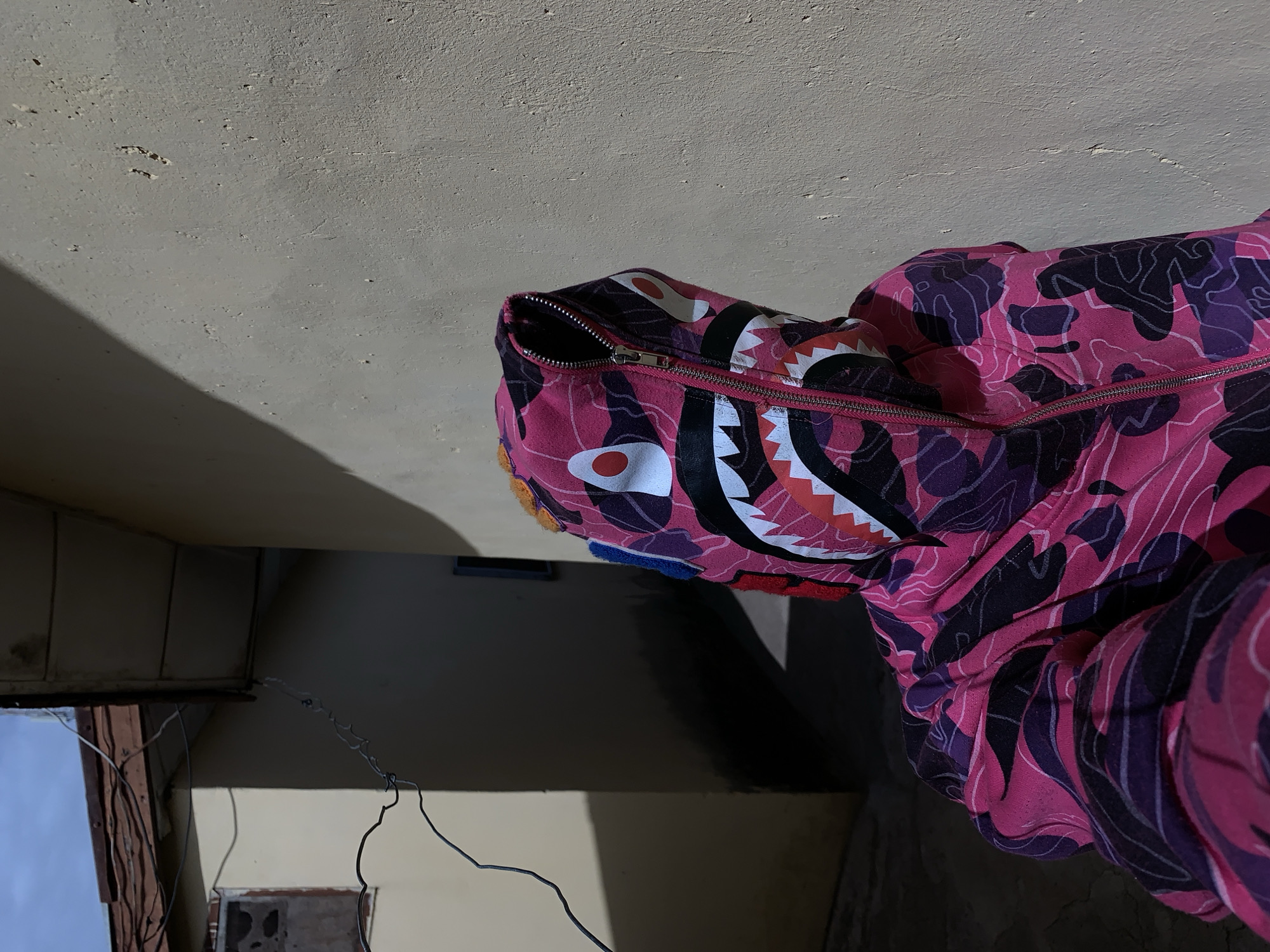 A Bathing Ape (BAPE) shark hoodie with a pink camouflage pattern is lying on a beige surface in an interior space. The hoodie is partially zipped and displays the shark face design on the hood. The environment appears to be a poorly lit room or hallway, with exposed wiring. No people are visible. This location is not readily identifiable in Rafin Par, Nigeria.