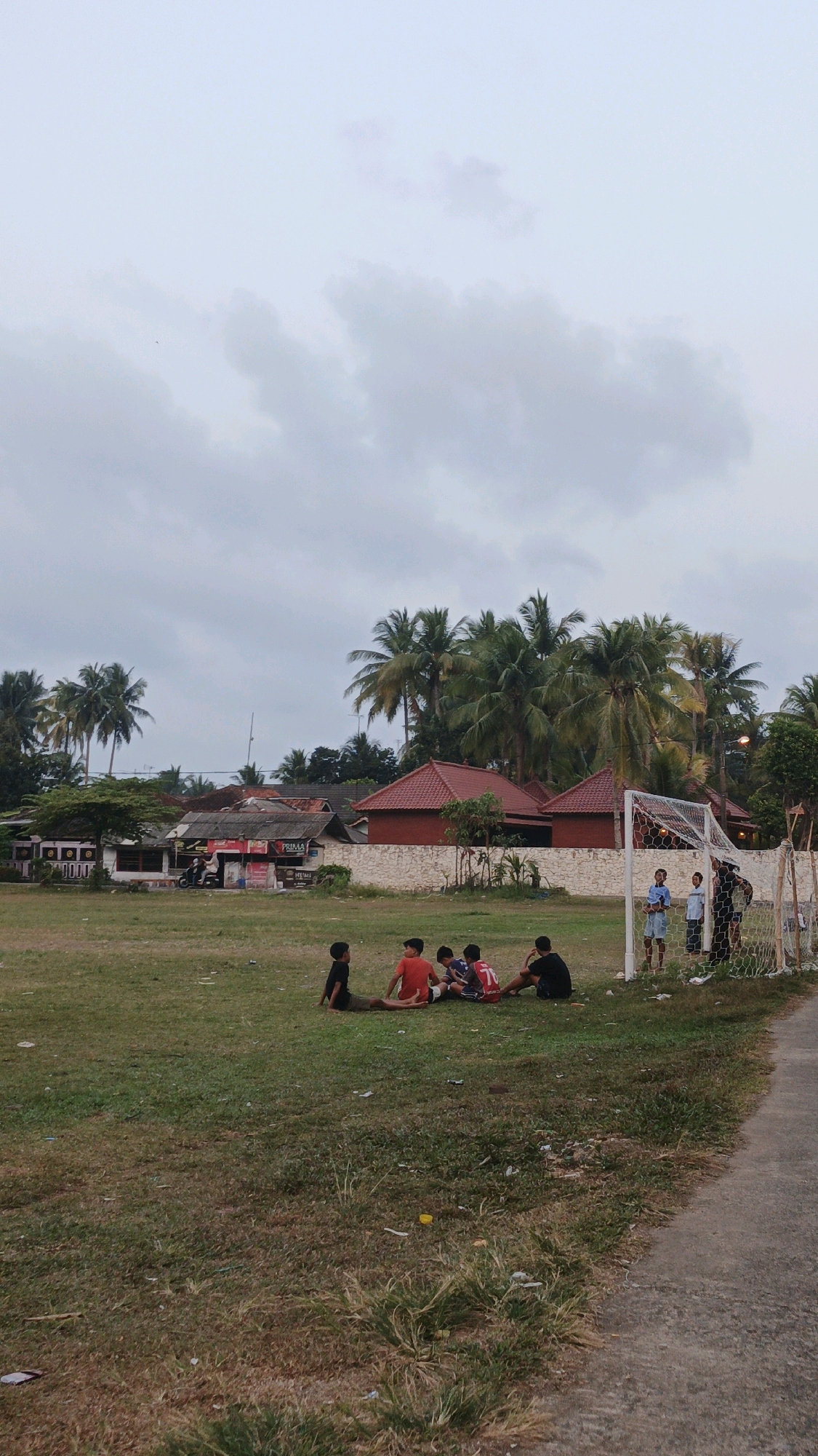 The image depicts a group of children playing soccer in a grassy field in Desa Cijulang, Indonesia. In the foreground, five boys are sitting on the grass near a soccer goal. Two of the boys are wearing red shirts, one is wearing a black shirt, one is wearing a white shirt with red trim, and another is wearing a red shirt with the number 71 on it. To the right of the goal, three other boys, dressed in blue shirts and shorts, and one person in a dark shirt and pants are standing near the goal. In the background, there are several buildings with red roofs and palm trees. The sky is overcast. A dirt road is visible on the right side of the image.