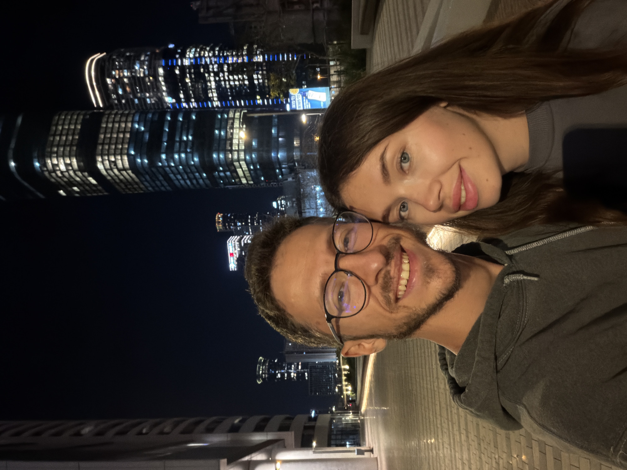A selfie taken at night in Tel Aviv, Israel, showing a man and a woman smiling at the camera. The man is wearing glasses and a grey hooded sweatshirt. The woman has long brown hair. Behind them, brightly lit skyscrapers create a dramatic urban backdrop. The foreground shows a paved pathway or street with reflective surfaces.