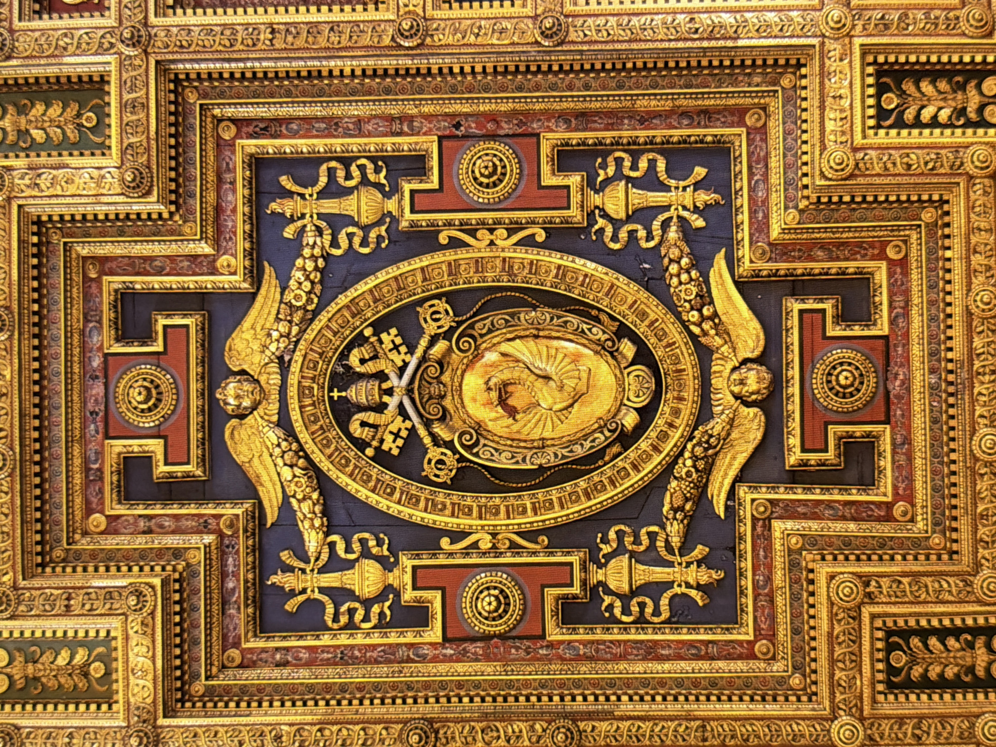 The image shows a richly decorated golden ceiling, likely an ornate interior. The ceiling is divided into geometric sections with intricate patterns, featuring motifs such as scrolls, floral designs, and what appear to be religious symbols like keys and a tiara. The central element is an oval frame containing an image of a winged figure, possibly an angel or a mythical creature, rendered in gold. This entire structure is heavily ornamented with gold leaf, creating a dazzling and luxurious appearance. The color palette is dominated by gold, deep blue, and red accents. There are no people or actions visible in the image, it is a static depiction of architectural detail.
This image does not depict a location in the city of Kyiv, Ukraine. The style and imagery suggest a European, possibly Italian, Renaissance or Baroque ecclesiastical or palatial interior.