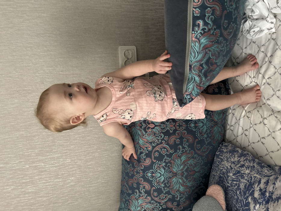 A toddler stands barefoot on a patterned bedsheet in an indoor room in Chaikovsky, Russia. The child has light-colored hair and wears a pink, ribbed two-piece pajama set with white cartoon bunny illustrations. The toddler's left hand rests on a large dark blue cushion patterned with intricate teal, light blue, and pinkish-brown paisley motifs. The right hand is positioned on a similar cushion to the child's right, which reveals a zipper along its edge. The child's gaze is directed off-camera to the right, with a slightly open mouth. The background features light beige textured wallpaper, and a white double electrical outlet is visible on the wall to the right of the child. Additional cushions, including one with a blue and white pattern and another grey and pink textured item, are visible on the bed to the left.