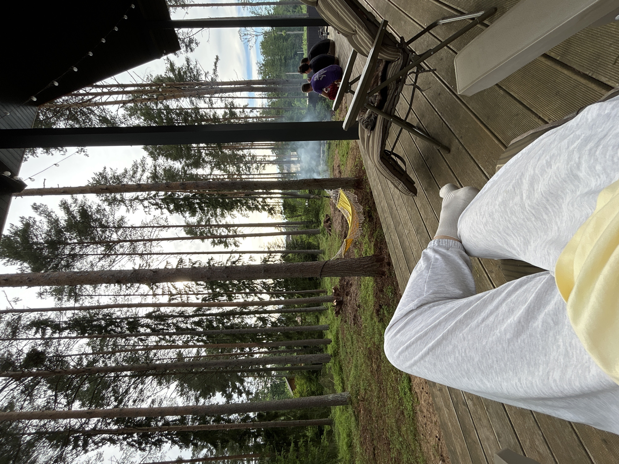 The image depicts a scenic view from a wooden deck in Shepelyovo, Russia. In the foreground, a person's legs clad in light grey sweatpants and white socks are visible, resting on the deck. A yellow garment is also partially visible. The deck overlooks a lush green forest with tall, slender pine trees. A yellow hammock is strung between two trees in the middle ground. Further in the background, a plume of smoke rises from a fire pit, suggesting a gathering or cooking activity. Several individuals are seated on chairs on the deck, observing the scene. The overall ambiance is one of relaxation and natural beauty.