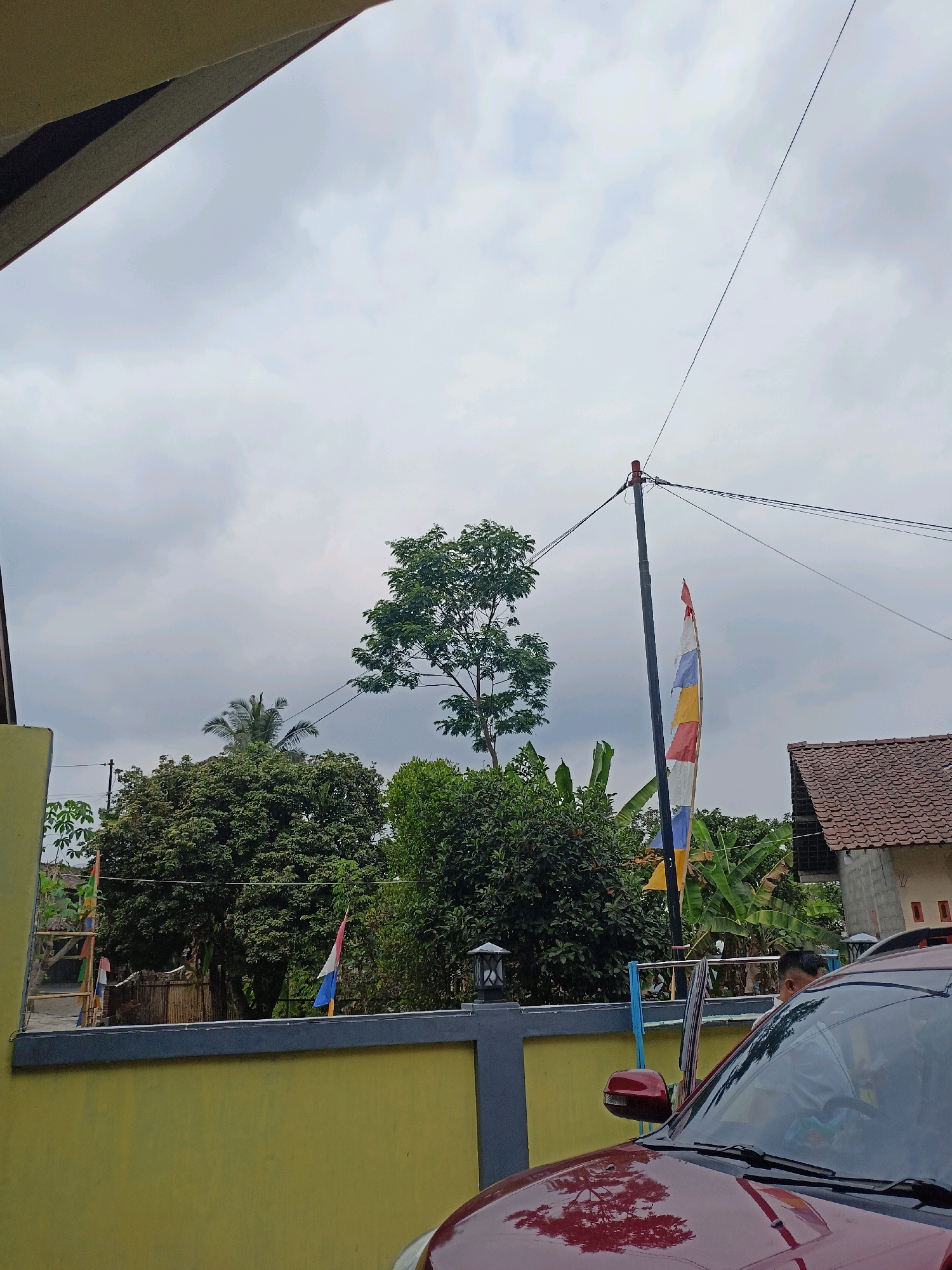 In Kalegen, Indonesia, a cloudy sky looms over a residential scene. A red car is parked in the foreground, its hood reflecting the surrounding environment. Behind the car, a yellow wall with a dark gray trim marks the boundary of a property. Beyond the wall, a variety of trees, some with lush green foliage, stand tall. A lone tree with a thinner canopy is situated towards the center of the frame. A utility pole with multiple wires stretches across the sky, and a tattered flag, displaying horizontal bands of red, white, blue, and yellow, hangs from it. To the right, a house with a terracotta tiled roof is visible. A person, partially obscured by the car and the open driver's side door, is seated inside the vehicle.