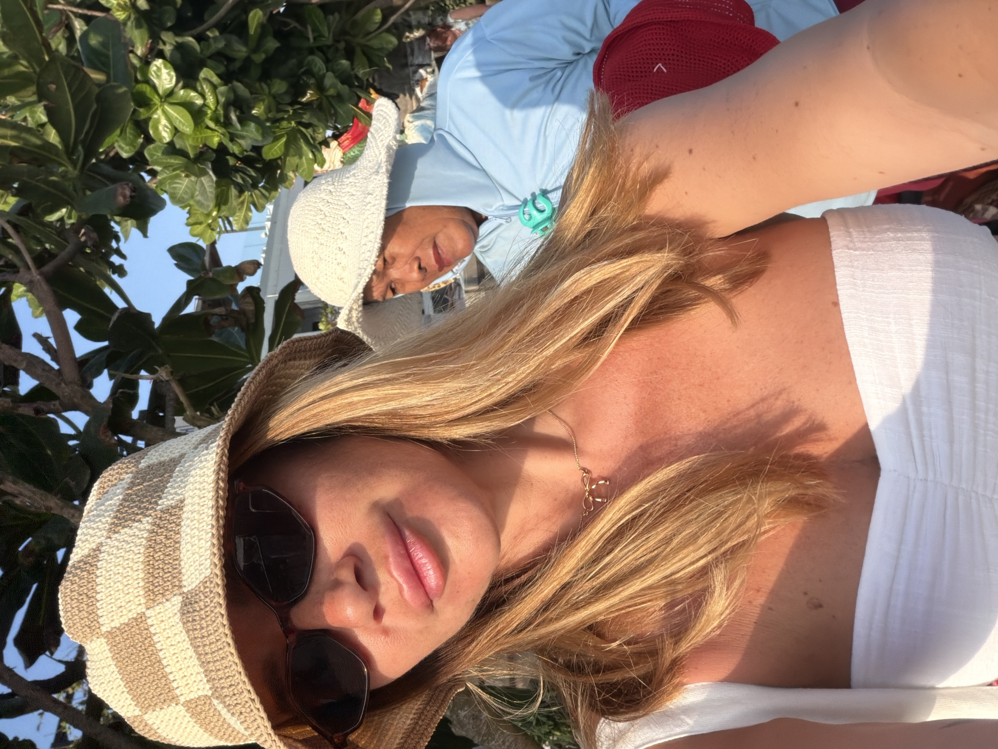 In a selfie taken outdoors with bright sunlight and foliage, a woman wearing a checkered bucket hat and sunglasses is in the foreground. She has blonde hair and is smiling, her lips slightly parted. She is wearing a white top and a delicate gold necklace. Behind her, another person wearing a white hat and a light blue hooded jacket is visible, partially obscured by foliage. The background suggests an outdoor setting with trees and possibly a beach. The image appears to have been taken in the city of Mengwi, Indonesia.