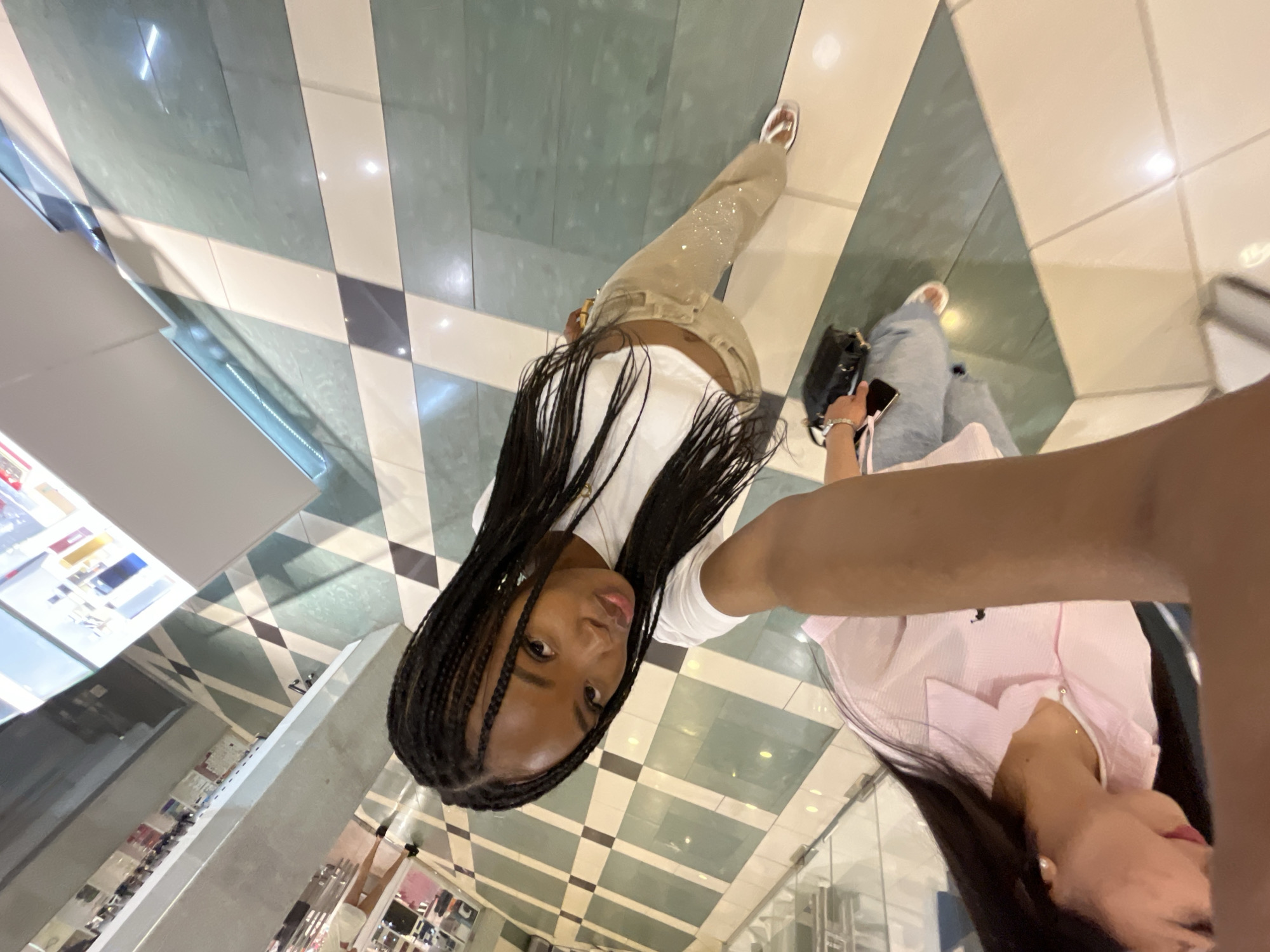 The image shows two young women in what appears to be a shopping mall. The woman in the foreground is taking a selfie, with her arm extended. She has long, braided black hair and is wearing a white crop top and beige pants with a sparkly pattern. The woman behind her is wearing a pink collared shirt. The background shows a tiled floor with a checkered pattern of white and green tiles, and reflective surfaces that suggest a well-lit interior space, possibly with displays of merchandise on shelves in the distance. The overall impression is of a casual shopping trip. The location is not specified as Kazan, Russia, and there is no information in the image to suggest this is the case.