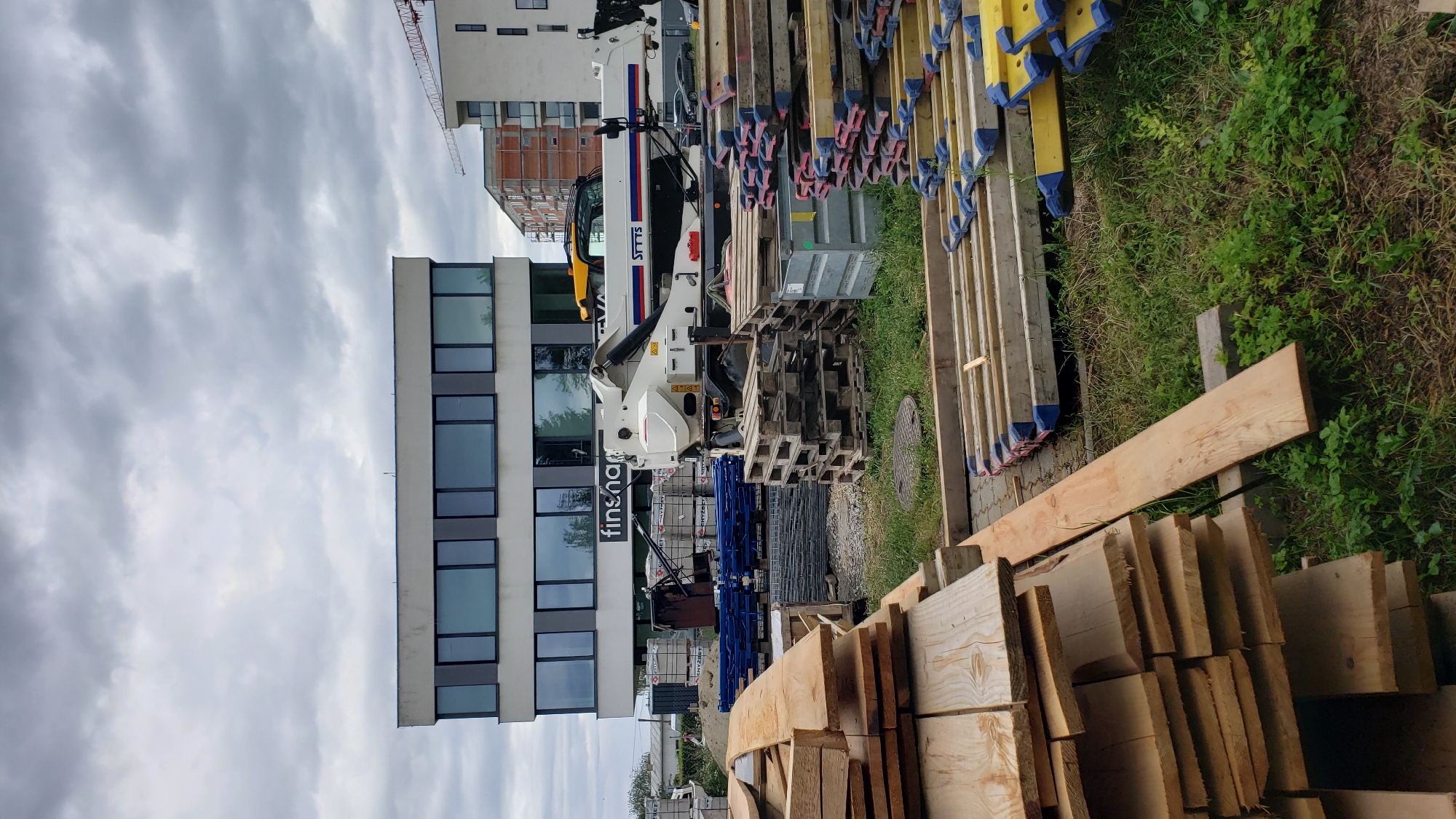The image depicts a construction site in Prešov, Slovakia, with a modern office building in the background and a large amount of building materials in the foreground. The office building features a sleek, contemporary design with large, reflective windows. A white construction crane is visible in the distance, indicating ongoing construction work. In the middle ground, a white truck with a crane arm is parked, and various stacks of lumber, wooden pallets, and metal scaffolding are organized on the ground. The overall scene is one of active construction and material storage, with a cloudy sky overhead.