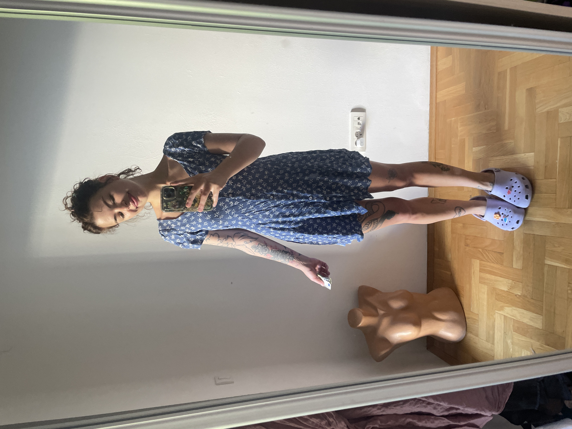 In Boreti, Montenegro, a young woman with curly brown hair is captured in a full-length mirror selfie. She is wearing a short, blue floral-print dress and light purple Crocs adorned with charms. Her arms are covered in tattoos, and she is holding a smartphone in her right hand, taking the picture, and a small white object, possibly a ticket or receipt, in her left hand. To her left, a beige torso mannequin stands on a light brown wooden floor. The wall behind her is white, and a power outlet with switches is visible on the right side. The room appears to be a domestic indoor space.