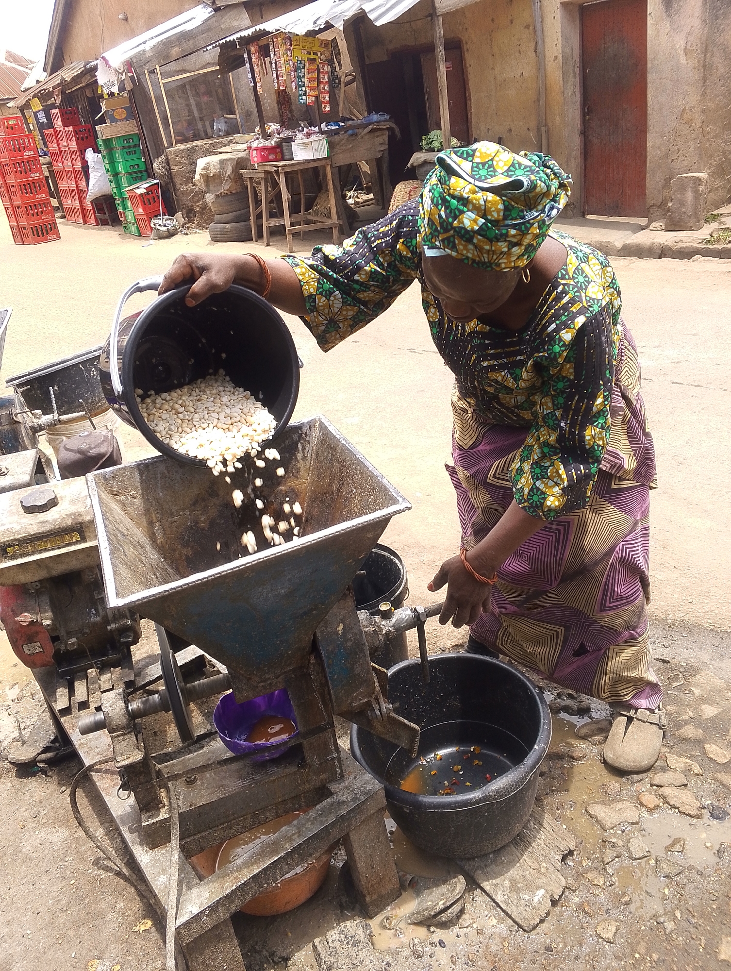 This image depicts an outdoor market or street scene in Jos, Nigeria, during daylight hours. The central focus is a woman, dressed in a vibrant green, yellow, and black patterned headwrap and matching traditional attire (a long-sleeved top and a patterned skirt/wrapper), engaged in food processing.
She is actively pouring white kernels, which appear to be maize or corn, from a black plastic bucket into the large, metal hopper of a grinding machine. The machine, possibly powered by an engine with "SUBAHUL" or "SUBAHULDEEN" visible on its casing, is processing the kernels into a thick, yellowish liquid or paste, which is being collected in another black plastic bucket placed beneath the machine's outlet. A smaller purple bowl and an orange terracotta bowl are also visible near the machine's base. The woman's right hand is near a lever or tap on the machine, suggesting she is operating or adjusting it.
The ground around the machine is unpaved, dusty, and somewhat muddy in places, with scattered small stones. In the background, several rustic market stalls or shops with corrugated iron roofs line the street. Piles of red and green plastic crates, many clearly labeled "Coca-Cola," are stacked against the buildings, indicating a commercial area. The scene portrays a typical everyday activity in a bustling local community.