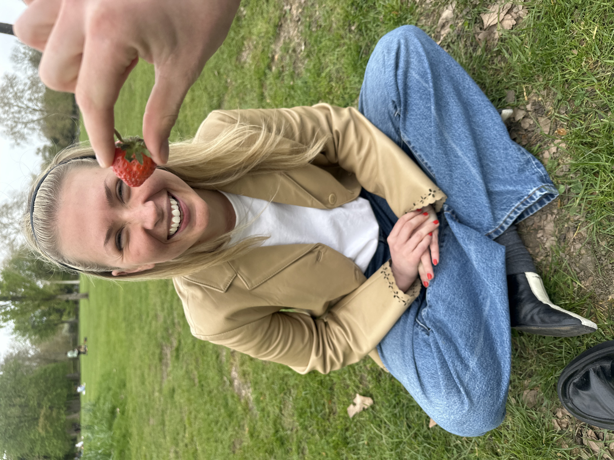 A woman with blonde hair and a black headband is sitting cross-legged on a grassy field. She is wearing a white t-shirt, a light brown jacket, and blue jeans. She is smiling up at a hand holding a strawberry over her head. She has red nail polish. A pair of black and white boots is visible next to her. The background consists of green grass and trees. This scene appears to be in a park-like setting, and given the general aesthetic, the park is likely located in the city of London, United Kingdom.