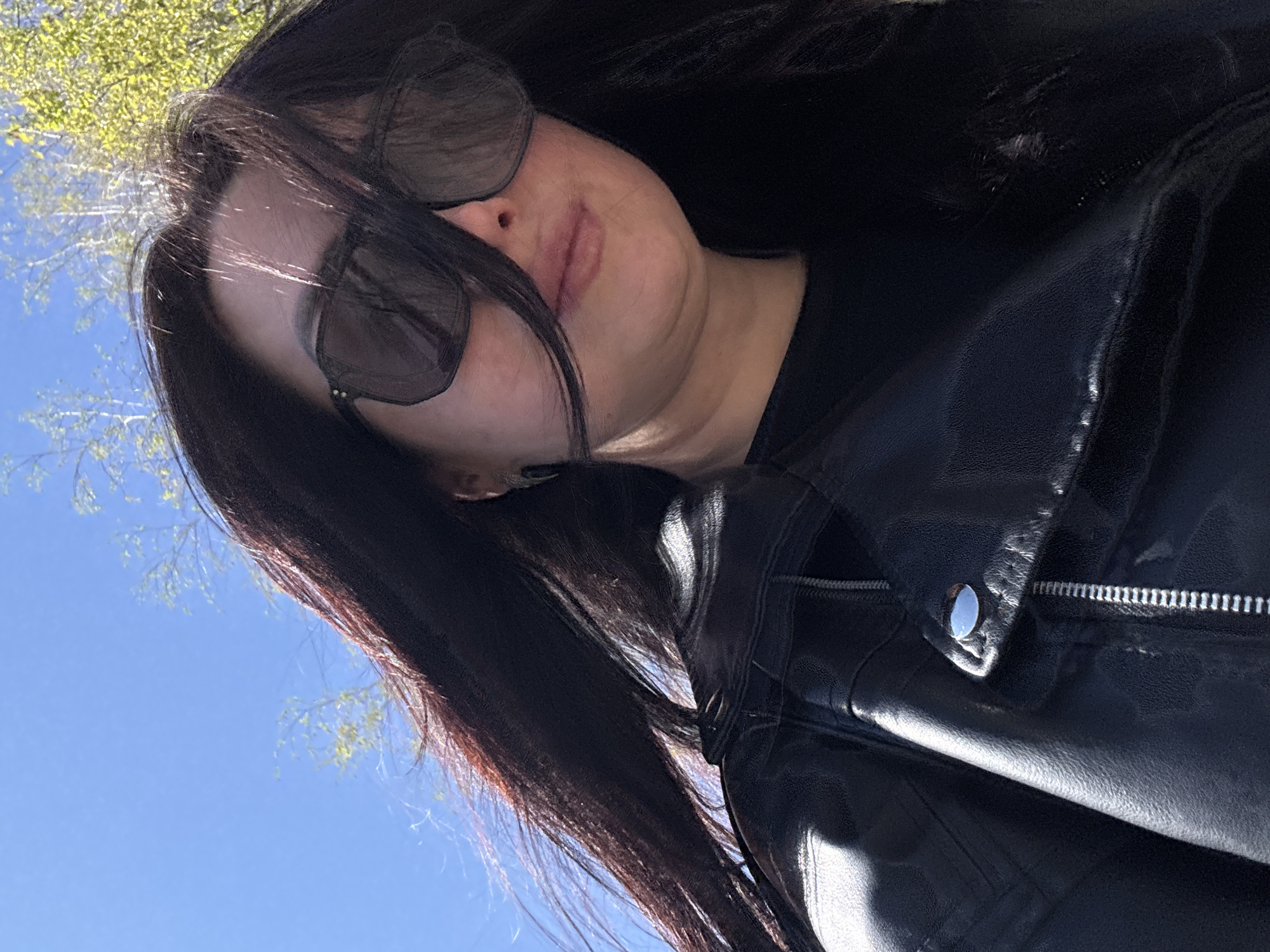 The image presents a person, likely female, captured from a low-angle perspective looking upwards. The individual has long, dark hair that partially obscures their face and is wearing dark sunglasses and a black leather jacket. The sunglasses cover the eyes, while a portion of the nose and lips are visible. The black leather jacket features a prominent silver zipper and a snap button detail near the collar.
The setting is outdoors on a bright, sunny day, evidenced by the clear blue sky and the reflection of sunlight on the jacket's surface. In the background, out-of-focus tree branches with sparse green leaves are visible against the vibrant blue sky, suggesting a spring or early summer season. The upward camera angle gives a worms-eye view, possibly indicating a selfie or a casual portrait taken from below. No specific activity is depicted beyond posing for the photograph. There is no visible text in the image.