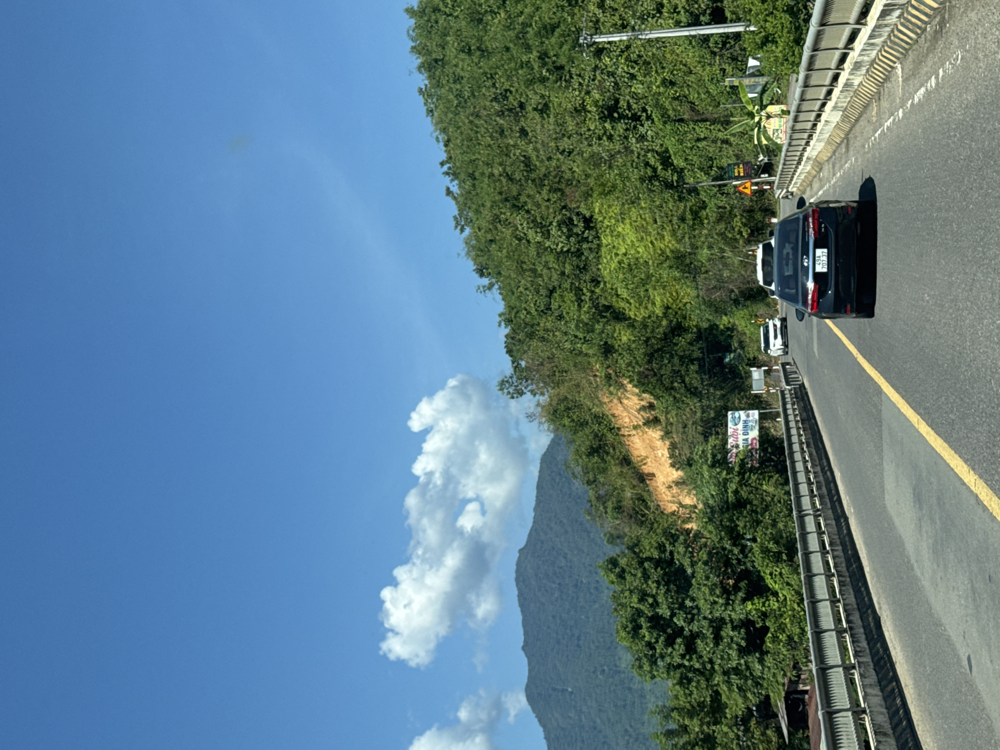 The image depicts a view from a vehicle driving on a bridge in Nam Khánh Vĩnh, Vietnam, on a clear day with a bright blue sky and scattered white clouds. The bridge has concrete railings with metal bars and yellow and black striped borders. The road is paved asphalt with a yellow lane divider.
Several cars are visible on the road. In the foreground, a black sedan with a license plate reading "49A 707.37" is driving away from the camera. Further ahead, a white SUV and a white sedan are also on the road.
The bridge spans across a vegetated area with lush green trees and a steep, orange-brown hillside. In the background, a large, tree-covered mountain dominates the landscape. A small, weathered wooden structure is partially visible on the left side of the road, near the bridge's edge. Signage is present along the roadside, including a white sign with Vietnamese text that appears to be advertising a business. A traffic sign with a yellow triangle and a warning symbol is also visible.