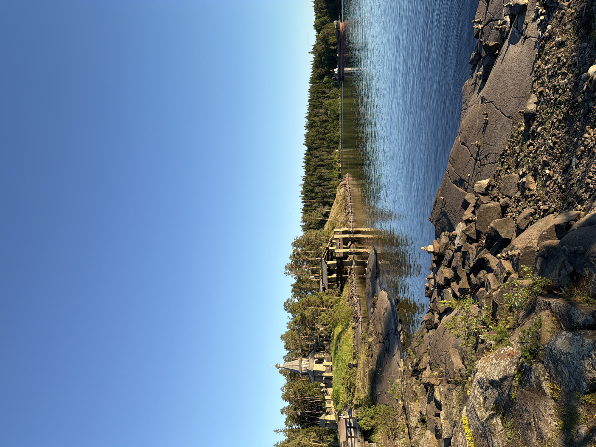 This is a landscape photograph taken on Valaam Island, Russia. The image depicts a serene waterscape with a clear blue sky. In the foreground, a rocky shoreline with scattered stones and small green plants is visible. The water is calm, reflecting the surrounding trees and sky.
On the left side of the image, a wooden walkway leads to a stone church with a grey, tiered roof topped with a golden dome. The church is surrounded by lush green grass and mature pine trees. A wooden structure, possibly a boathouse or pier, is situated on the right side of the church, also built with stone and wood. A large barge or ship is seen in the distance on the right, navigating the water.
The overall scene conveys a sense of tranquility and natural beauty, characteristic of the Valaam archipelago. The lighting suggests it is either early morning or late afternoon, with the sun casting long shadows.